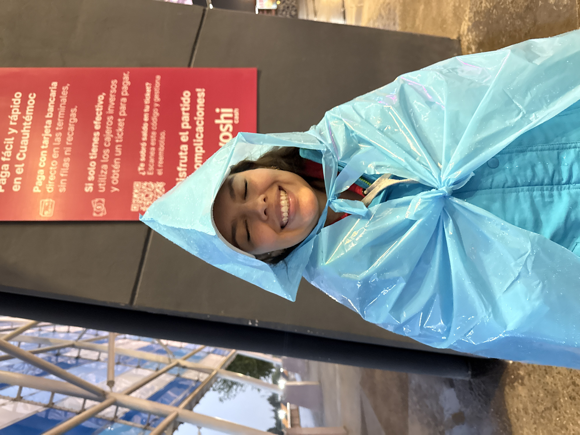 A woman wearing a blue poncho smiles at the camera. Behind her is a red sign that says "Paga fácil y rápido en el Cuauhtémoc," indicating that she is at the Estadio Cuauhtémoc in Puebla, Mexico. The sign provides information about payment options for tickets. A metal structure is also partially visible in the foreground.