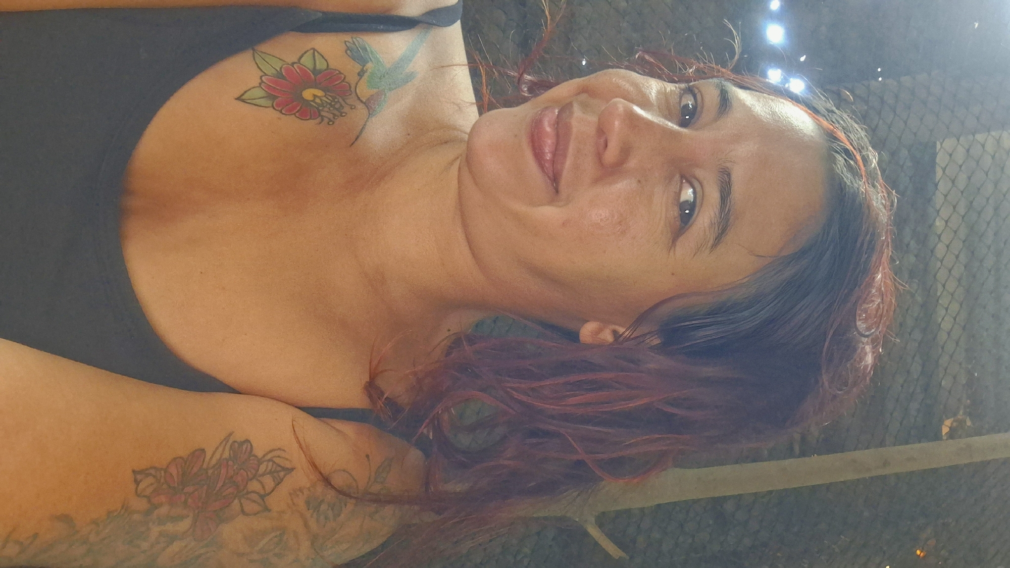 A woman with reddish-brown hair tied back poses for a selfie in a dimly lit, outdoor setting, possibly in Vallecito, Venezuela. She is wearing a dark tank top and has tattoos on her chest, including a hummingbird and a flower, and on her right arm with floral designs. The background shows a dark, textured surface, possibly a fence or netting, with some blurry lights in the upper left corner and a vertical wooden pole on the right. Her expression is a slight smile, with her tongue peeking out slightly.