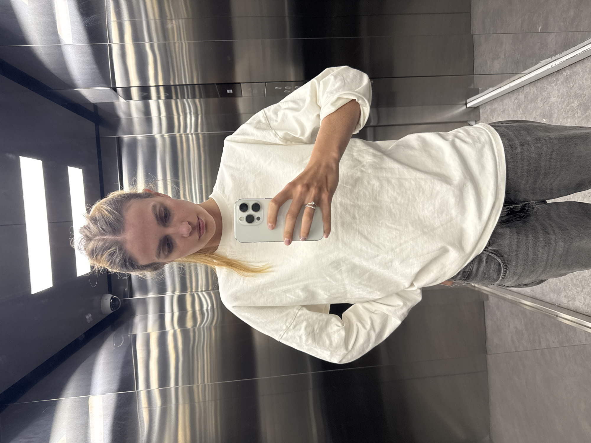 A woman is taking a selfie in an elevator. She is wearing a white long-sleeved shirt and gray jeans. She is holding a white smartphone in her right hand. The elevator has stainless steel walls and a dark gray floor. The location is Unknown, Unknown.