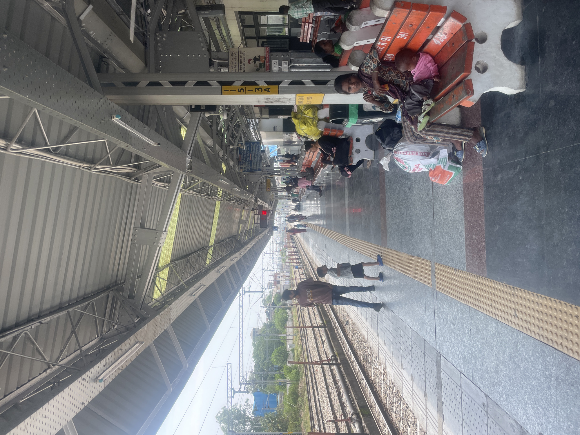 The image shows a view of a covered train platform at Pathankot Junction railway station in Pathankot, India. The platform is paved with grey tiles and features a yellow tactile paving strip for visually impaired passengers. Numerous people are visible on the platform, some sitting on benches, others standing or walking. One man and a young boy are standing near the tracks, looking towards the distance. A woman and a baby are seated on an orange and white bench. Luggage is present on the platform, including bags and a large yellow object wrapped in plastic. The platform is covered by a corrugated metal roof supported by a steel structure. Overhead wires and railway tracks are visible in the background, along with trees and buildings. Signs with text in Hindi and English are posted on pillars. The overall scene depicts a typical bustling Indian railway station environment.