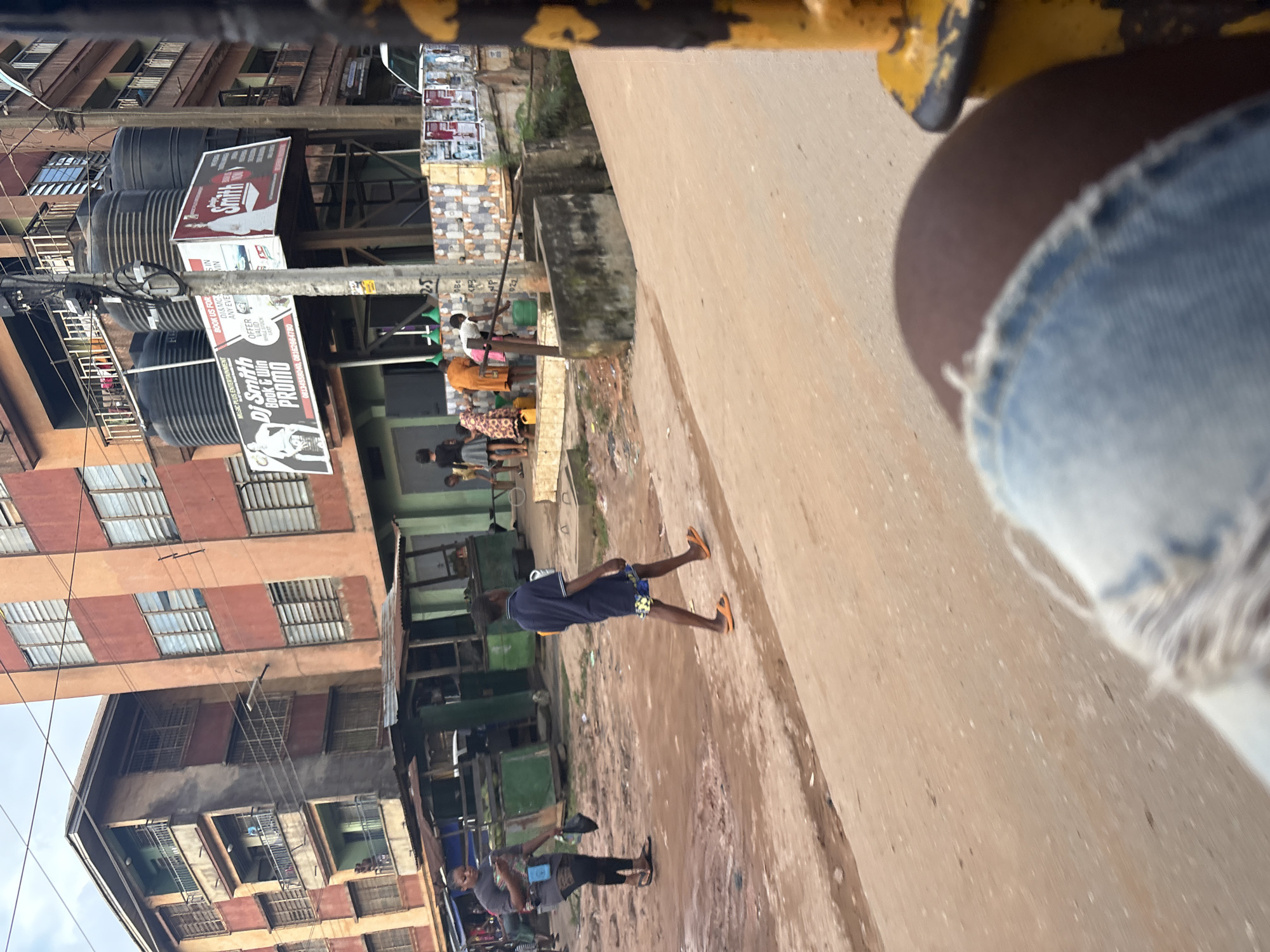 The image captures a street scene in Onitsha, Nigeria. A young boy in a dark blue shirt and patterned shorts walks across a dirt road, his orange flip-flops visible. To his left, a woman in a gray t-shirt and black leggings stands, holding a dark bag and looking at her phone. In the background, a multi-story building with balconies and numerous windows is prominent. Several market stalls with green awnings are set up along the street. A group of people, including children, are gathered around a water source where someone is filling a yellow container. Large black water tanks are mounted on a platform above the street, and promotional posters are visible on the building's facade. The environment appears dusty and the sky is overcast. The foreground is partially obscured by the leg and denim shorts of the photographer, and a yellow pole with black markings is visible on the right side of the frame.