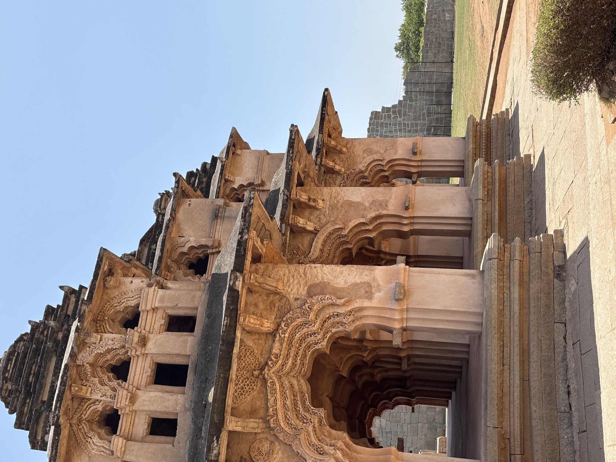 This outdoor, medium-angle shot features the Lotus Mahal building located in Kamalapura, India. The building is constructed from a tan-colored stone. The perspective is looking up at the side and roof of the building. The roof has a series of triangular elements that are stacked on top of each other. The side of the building features a repetitive archway design. The arches are intricately carved with floral and geometric designs. The building has windows. A stone wall with stone stairs on the top right part of the frame. Above the wall is grass, trees, and blue sky. The light is bright and diffused.