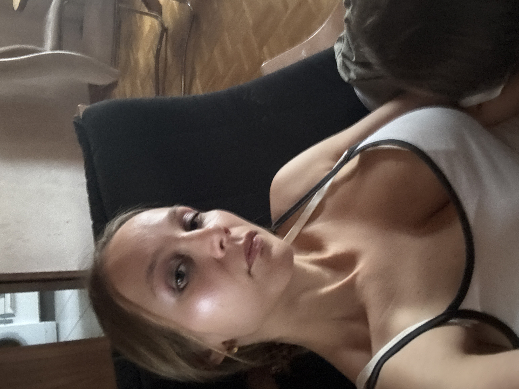 A young woman with blonde hair is sitting in a chair, holding a baby. The woman is wearing a white tank top with black trim. The baby's head is partially visible, nestled against the woman's chest. The background shows a room with wooden floors and furniture. The lighting suggests it is daytime.