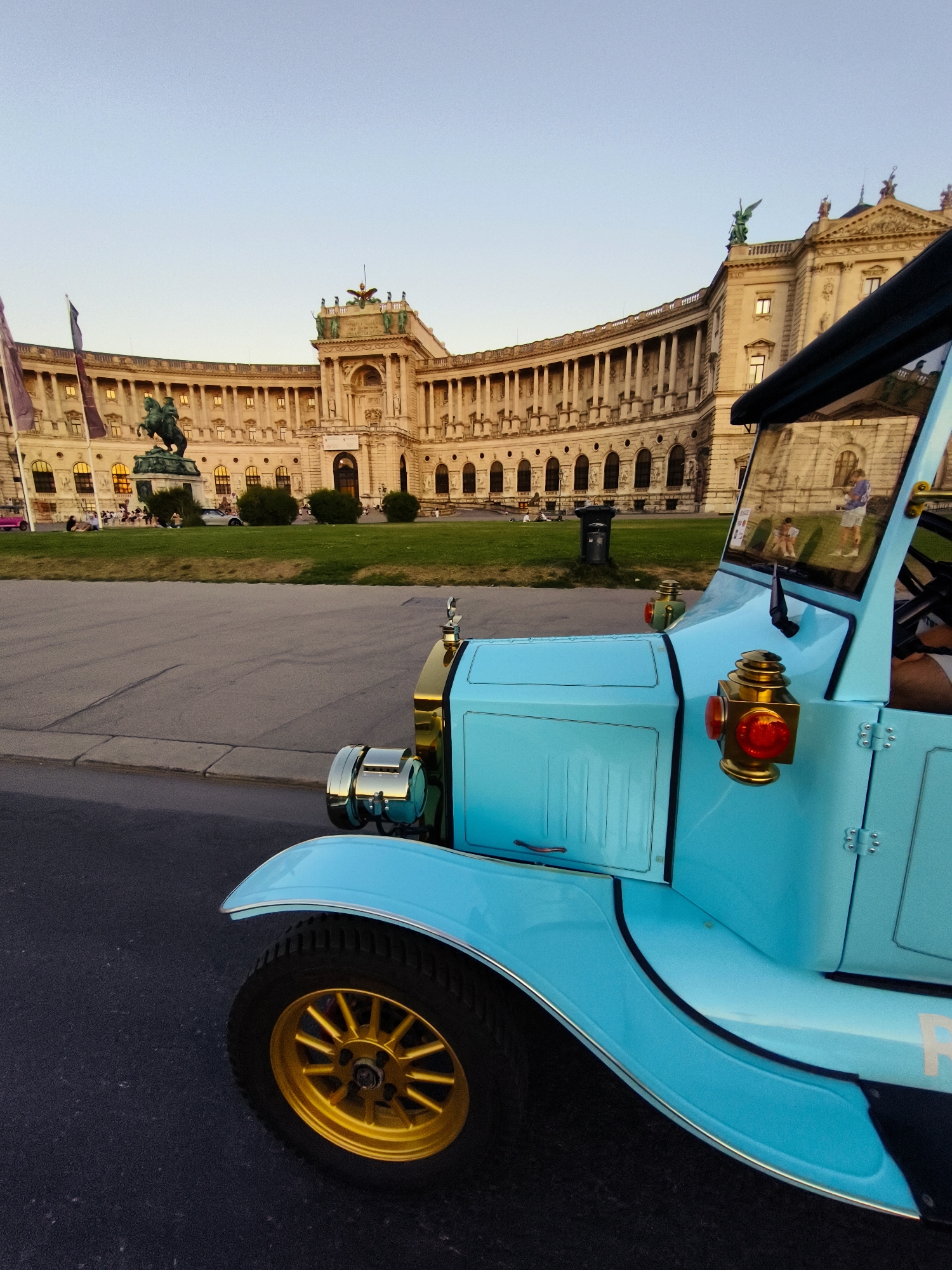 In Vienna, Austria, a bright blue vintage car with gold accents is parked on a paved street in front of the Hofburg Palace, a grand imperial palace. The car is positioned at an angle, with its front right wheel and fender prominently featured. The Hofburg Palace, with its distinctive arcaded facade and ornate architecture, serves as a backdrop. The palace grounds include a manicured lawn and trees, and a statue of a man on horseback is visible. The sky is clear and blue, suggesting a daytime scene, possibly late afternoon due to the warm light. Several people are seated on the grass in the distance, and a few individuals are seen walking near the palace entrance. The overall atmosphere is serene and picturesque, capturing a moment of historical grandeur combined with a playful, retro element.