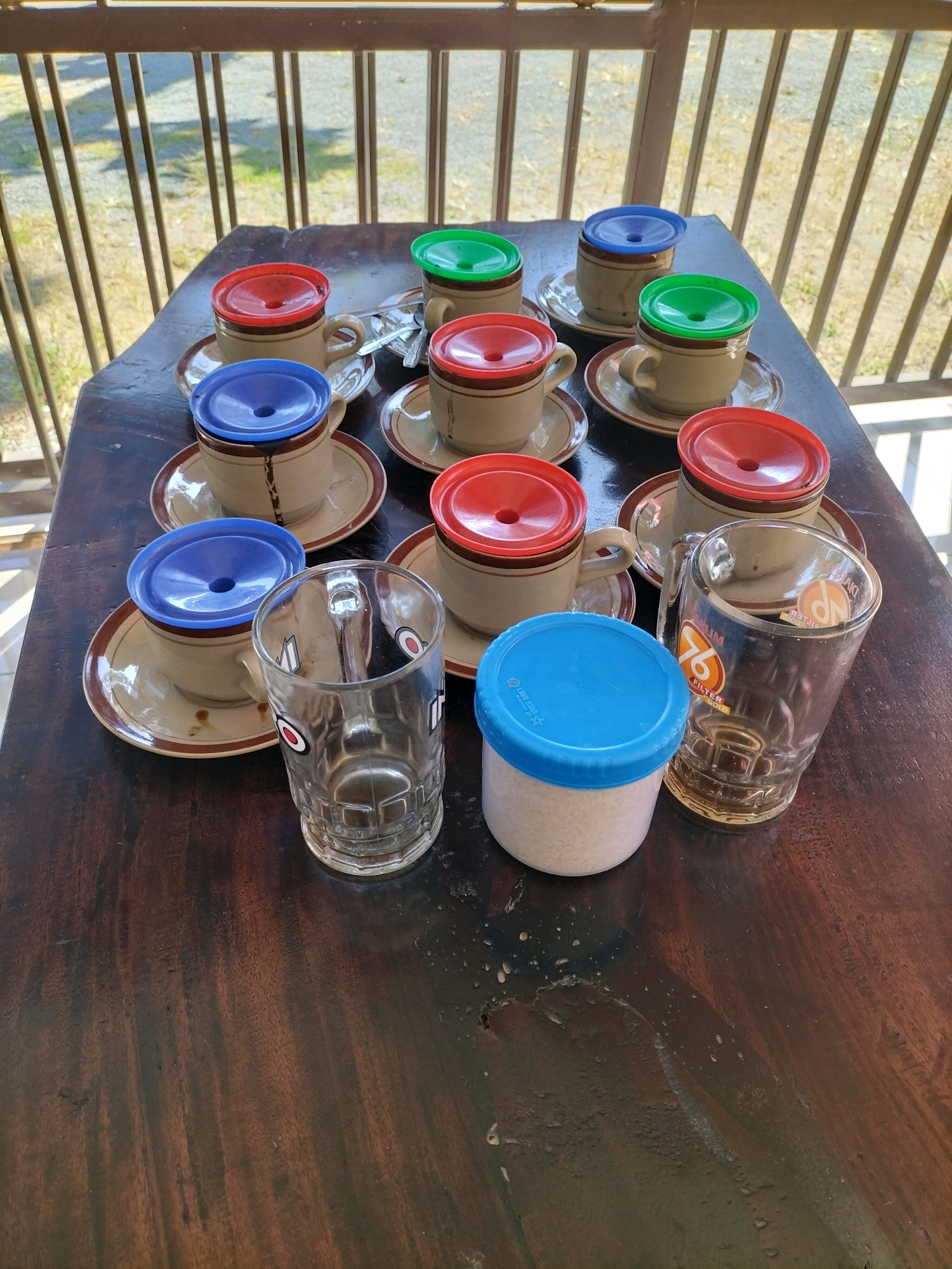 The image depicts a collection of coffee cups and saucers arranged on a dark wooden table, likely outdoors due to the presence of a railing and natural light filtering through. There are at least ten coffee cups visible, each with a saucer. Several cups are covered with brightly colored plastic lids: red, green, and blue. Some of the cups have handles. In addition to the cups, there are two clear glass mugs on the table. One of the mugs is empty and has a label with text and a red and white circular logo. Next to it is a small white plastic container with a blue lid. The overall scene suggests a casual setting, possibly a cafe or a personal gathering, in Tasikmadu, Indonesia. The lighting suggests it is daytime.