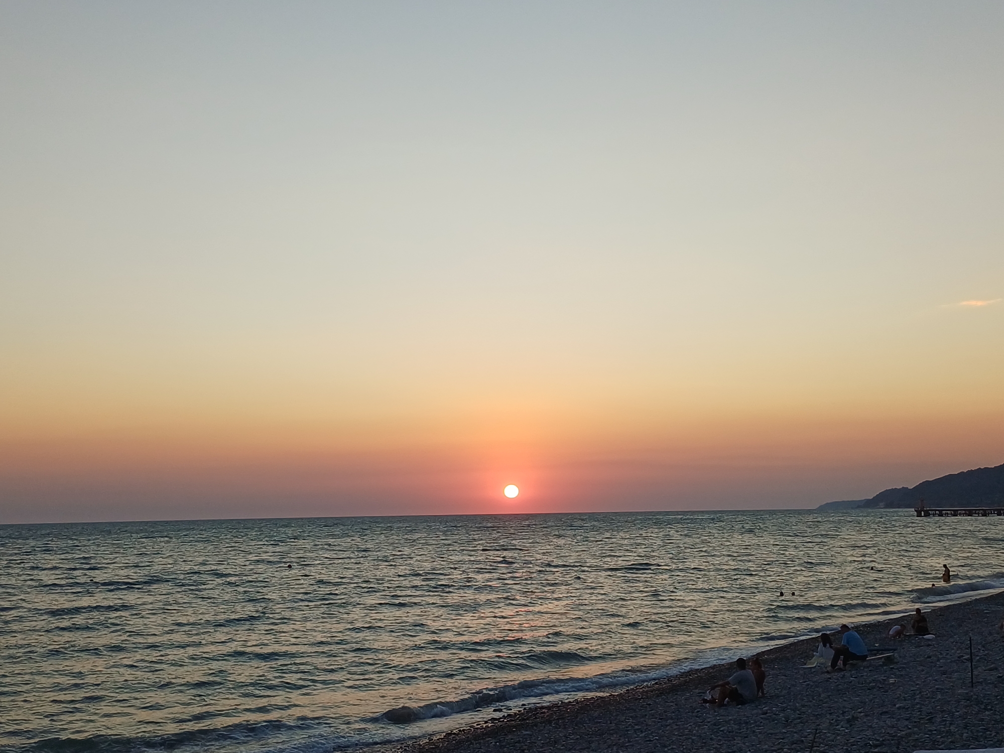 The image depicts a sunset over the Black Sea on a pebble beach in Sochi, Russia. The sun, a bright orb, is low on the horizon, casting a warm orange glow across the sky and reflecting on the water's surface. The sea is calm, with gentle waves lapping at the shore. Several individuals are visible on the beach, some sitting and others standing, enjoying the view. In the distance, a pier extends into the water, and a hilly coastline frames the scene. The overall atmosphere is serene and peaceful.