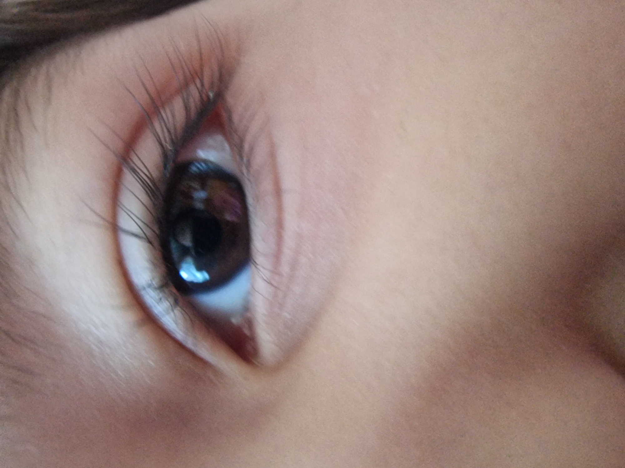 A close-up shot of a human eye. The eye has long, dark eyelashes. The iris is dark brown, reflecting a light source. The skin surrounding the eye is light-toned. No city is discernible from the image.