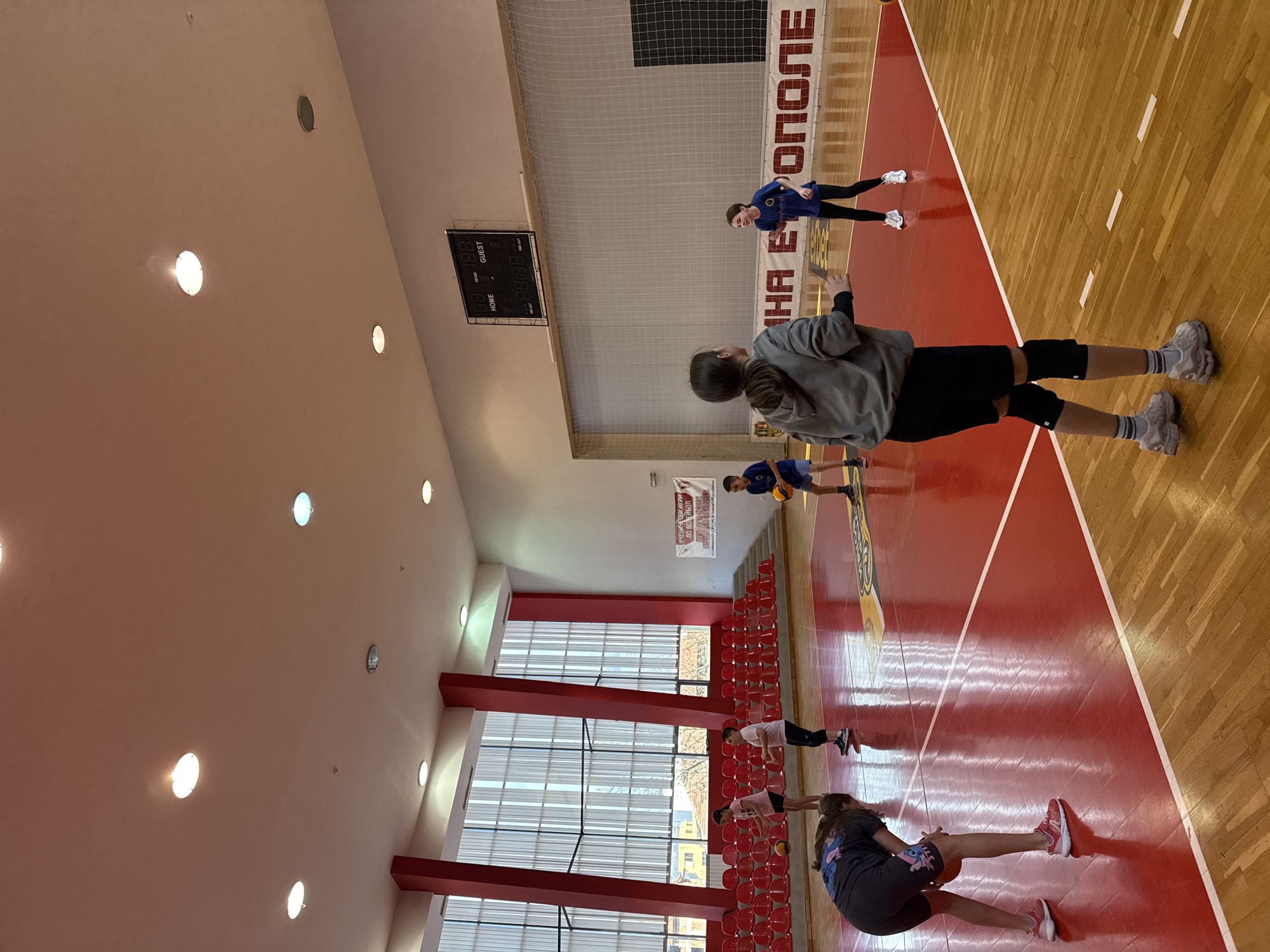 The image shows an indoor gymnasium in Etropole, Bulgaria, with a group of young people engaged in physical activity.
Several individuals are present on the wooden and red-painted floor of the gym. A girl in a gray sweatshirt, black shorts, knee pads, and gray sneakers is closest to the camera. Other individuals are wearing athletic attire, and some are stretching or performing exercises.
A digital scoreboard is visible on the wall, along with signage in Cyrillic. Windows run along one side of the gym, with red structural supports and rows of red seating beneath. The ceiling is white with recessed lighting.