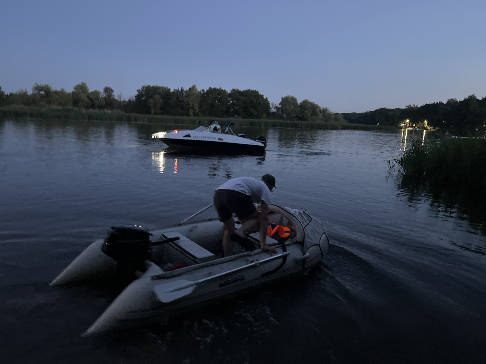 An inflatable boat with an outboard motor is in the foreground on a body of water. A person in a white t-shirt and dark shorts is leaning into the inflatable boat, and an orange life vest is visible inside the boat. Further in the background, a white speed boat with an outboard motor is moving on the water. The sky is dark blue, indicating dusk or night. Trees and reeds line the distant shore. The scene appears to be a natural setting, likely a river or lake, possibly on the outskirts of Saratov, Russia, given the time of day and the presence of boats.