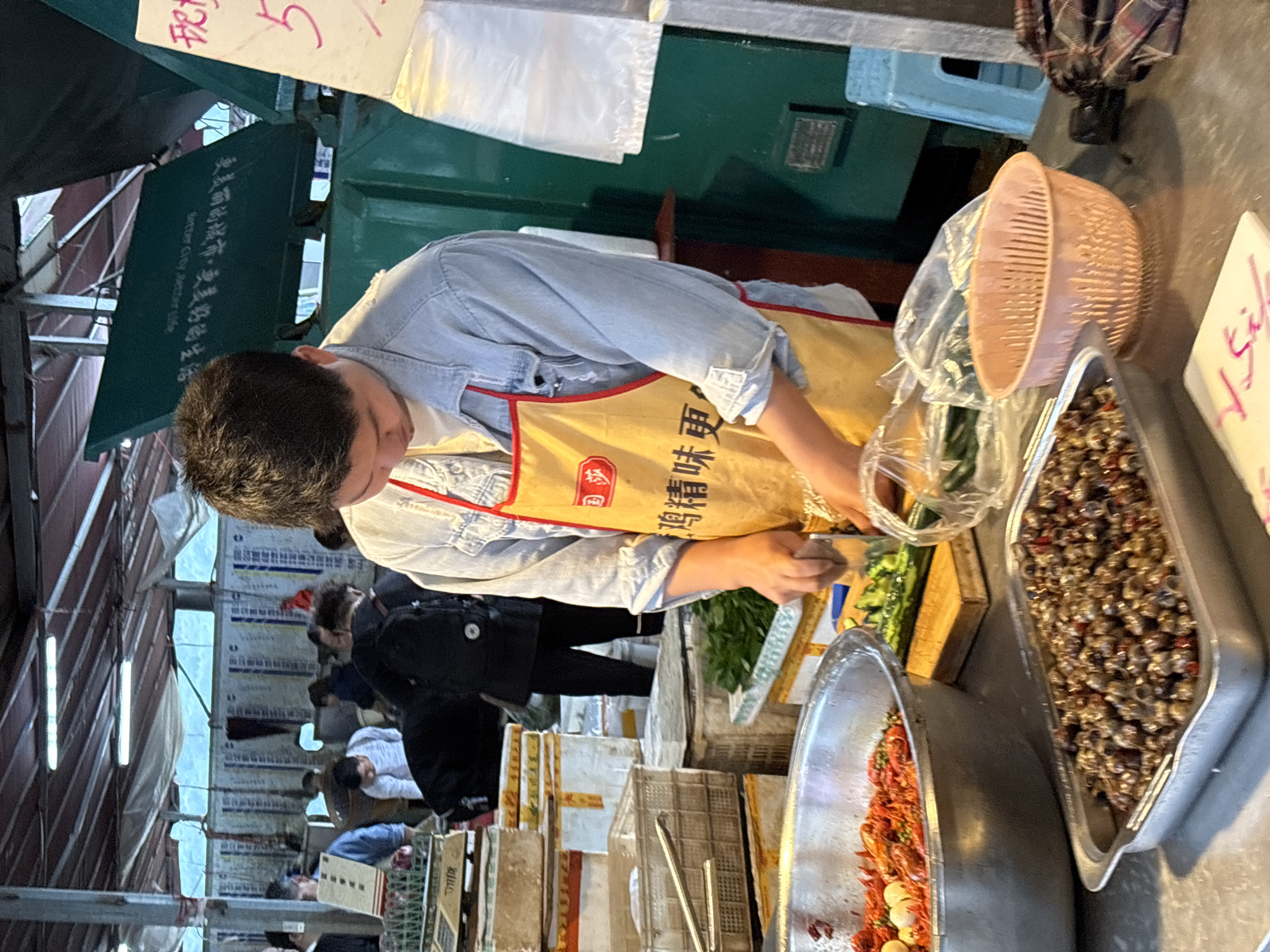 A street vendor in Jiangjin, China, wearing a yellow apron with Chinese characters, prepares food at a stall. He chops vegetables on a wooden cutting board next to trays containing cooked shellfish and chili peppers. Shoppers walk past the stall under a canopy of corrugated metal. The stall includes covered food displays and a pink plastic basket.