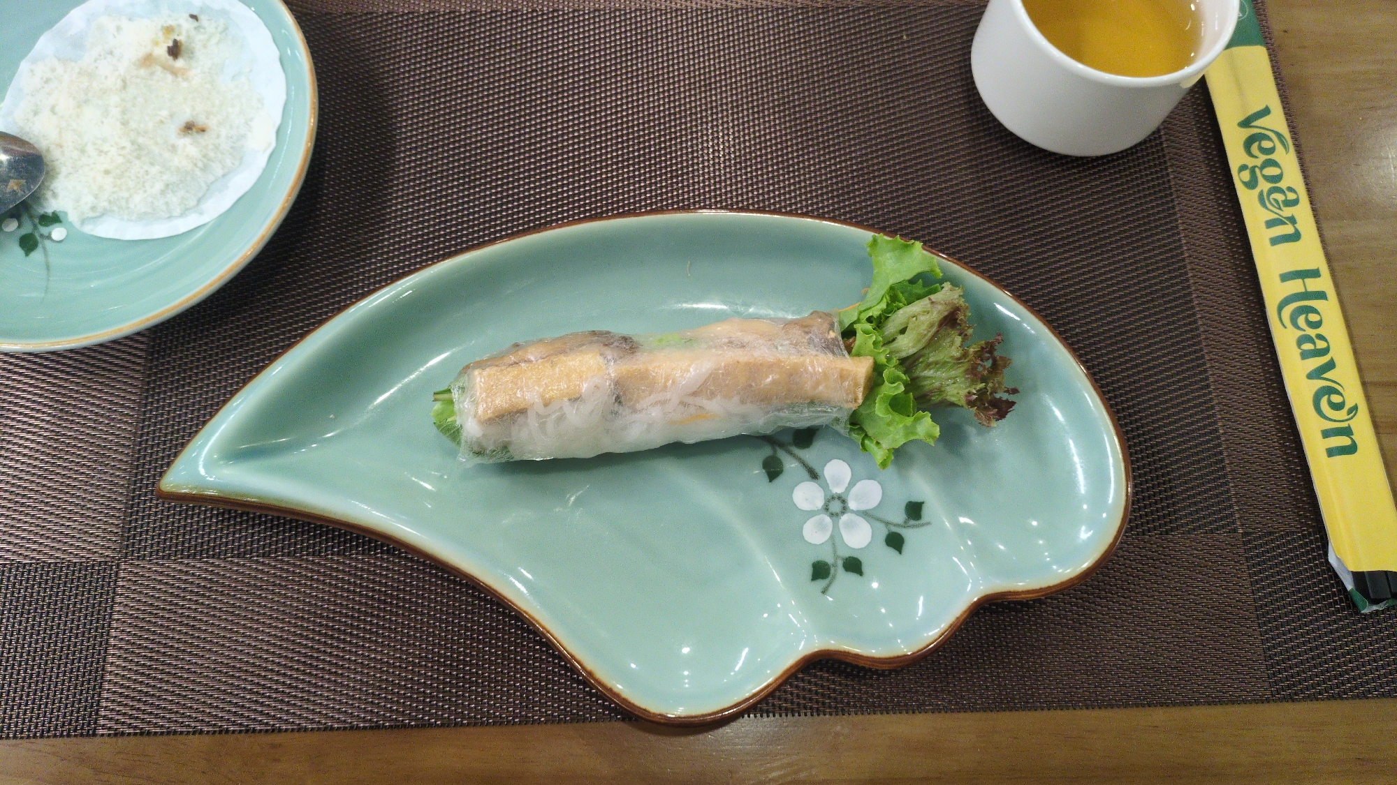 The image shows a table setting in a restaurant, likely "Vegan Heaven" based on the visible chopsticks. A fresh spring roll sits on a decorative, light green plate. Next to it is a pair of yellow chopsticks labeled "Vegan Heaven." A cup of liquid, possibly tea, is adjacent to the chopsticks. In the upper left corner, another plate holds a portion of a white, granular food item. The table surface is covered with a brown textured placemat. The setting suggests a meal in Da Lat, Vietnam.