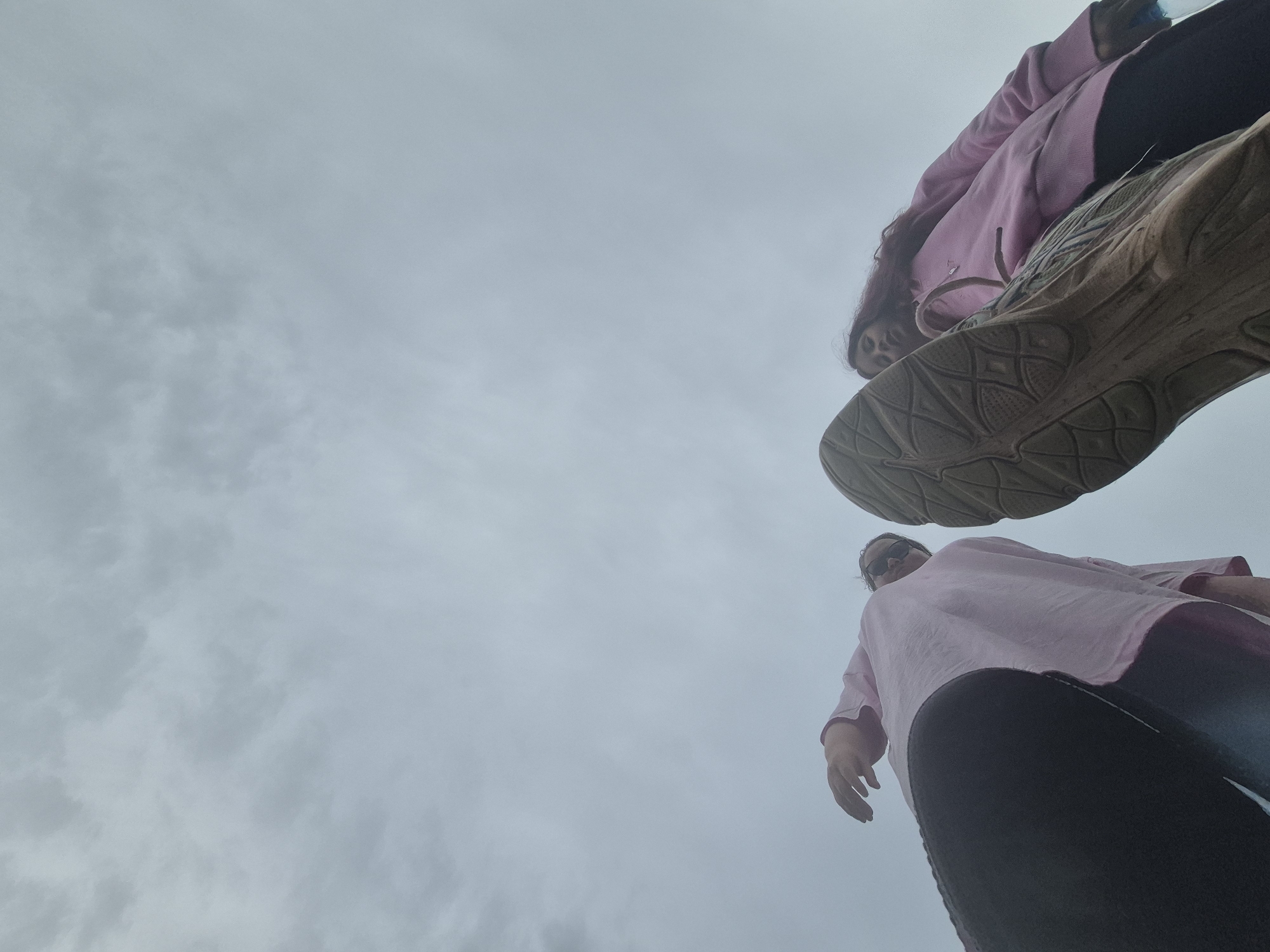 This low-angle photograph, taken from the ground looking upwards, depicts two individuals against an overcast sky. The camera's extreme perspective creates a "worm's-eye view."
In the upper right portion of the frame, the sole of a large athletic shoe, likely a sneaker, is prominently featured, displaying its intricate tread pattern. This shoe belongs to one of the subjects, who is seen wearing a pink long-sleeved top and dark pants or leggings. Her long, dark hair is visible, falling over her shoulder, and she holds a blue object, possibly a water bottle, near her chest. Her face is partially visible as she looks down towards the camera.
Positioned in the lower right, the second individual is seen from approximately the waist up. They are also dressed in a pink long-sleeved shirt and dark pants. This person appears to be wearing sunglasses, obscuring their eyes, and their right hand is visible. The edge of another shoe sole is partially visible above and to the right of this person's head.
The background is entirely composed of a uniformly grey, cloudy sky, suggesting overcast weather conditions during daylight hours. The two subjects appear to be standing directly over the camera, looking down, possibly engaging in a playful pose or taking a unique selfie. There is no discernible text within the image. The setting is Chaikovsky, Russia.