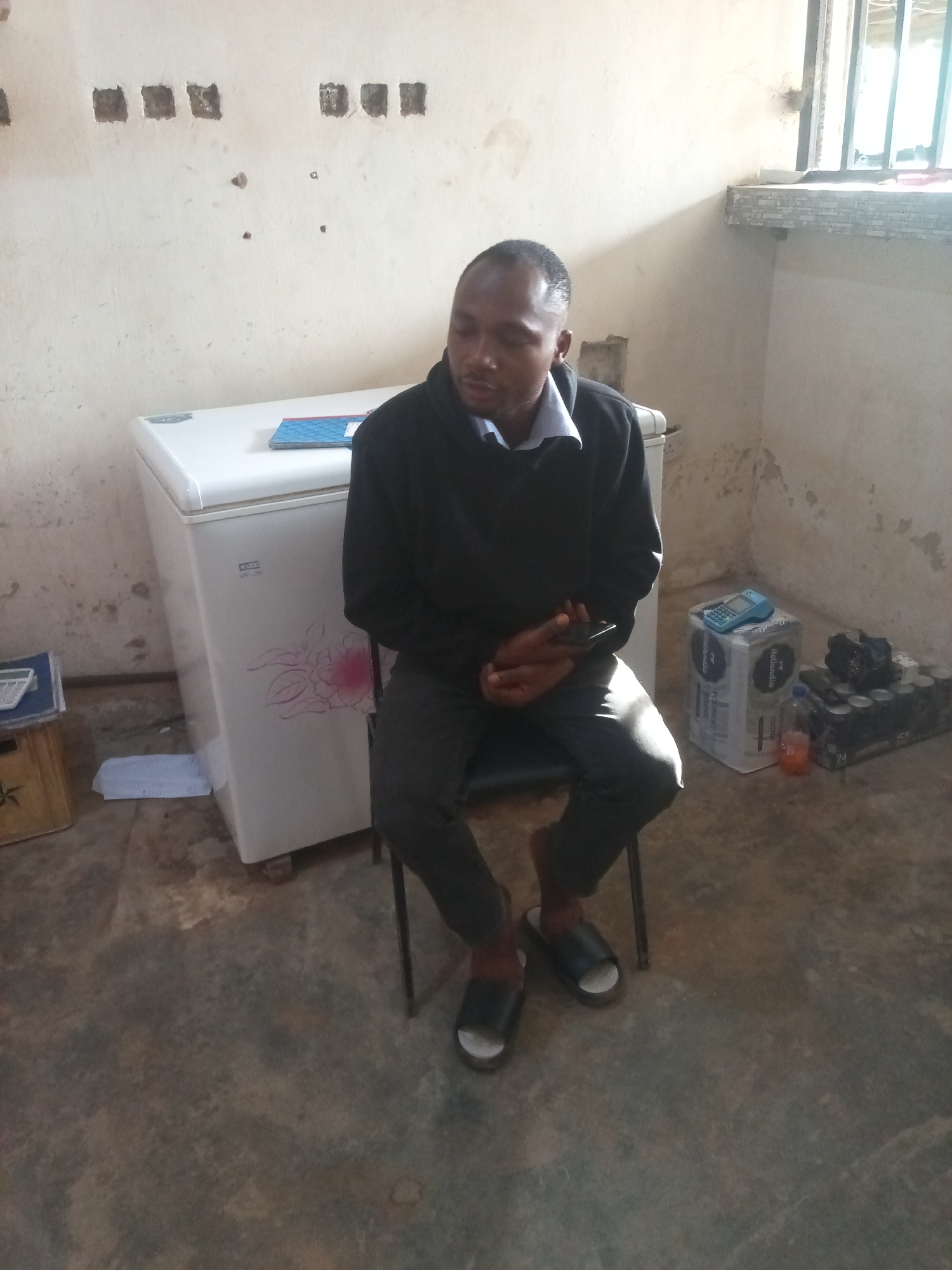 A Black man, wearing a dark-colored hooded sweatshirt and dark pants, sits on a black chair. He is holding a mobile phone in his hands. The man is seated in what appears to be a room with a light-colored, textured wall and a concrete floor. To his left is a white upright freezer with a pink floral decal on its side. On top of the freezer is a blue folder. To the right of the man, on the floor, is a stack of white boxes, one of which has the word "Hollandia" printed on it, and a blue calculator on top. Next to the boxes is a case of canned beverages. A window with metal bars is visible in the background, with light filtering through it. The overall environment suggests an indoor setting, possibly a storage area or a sparsely furnished room.