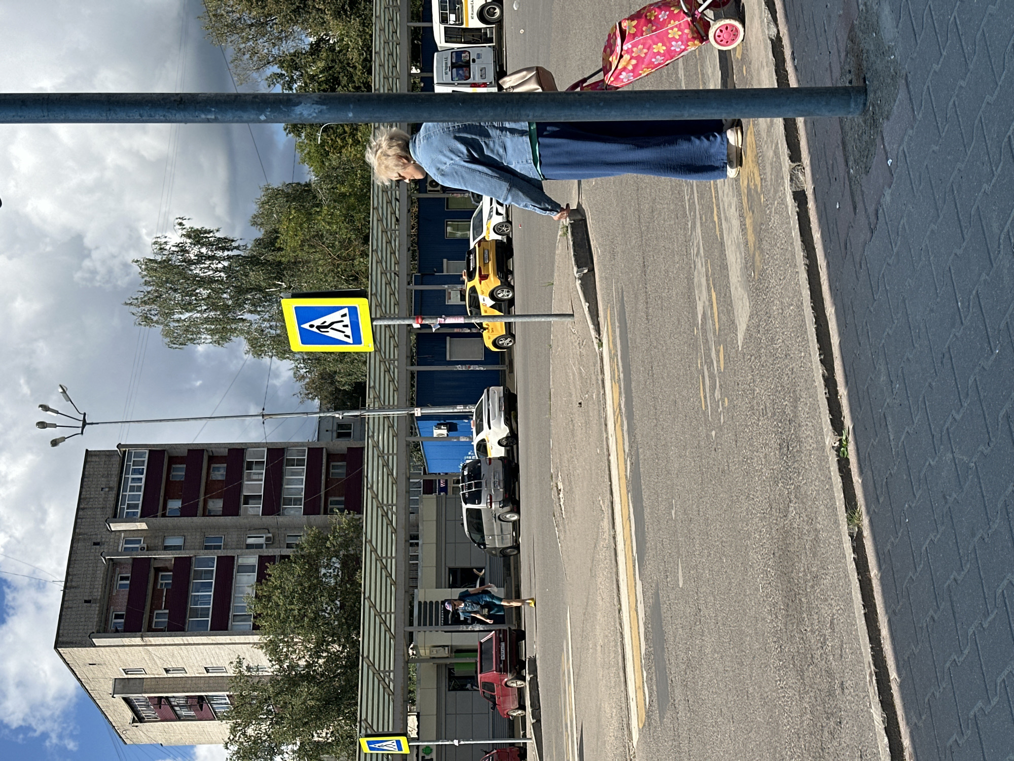 In Chekhov, Russia, on a cloudy day, a woman stands on a sidewalk next to a pedestrian crossing, holding the handle of a pink wheeled shopping bag with a floral pattern. She is wearing a denim jacket, a blue long skirt, and white shoes. To her left, across the street, a yellow taxi and a white car are parked near a covered bus stop. Further in the background, a tall, multi-story residential building with a light-colored facade and dark red balconies stands behind trees. A pedestrian crossing sign with a yellow background and a silhouette of a person walking is visible near a lamppost. The scene is set on a paved road with a marked pedestrian crossing.