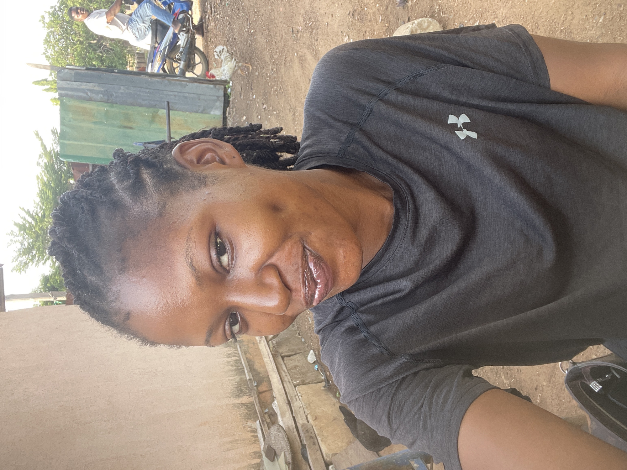 A medium, close-up shot, partially rotated, shows a Black woman wearing a gray Under Armour t-shirt. Her hair is styled in small braids. The environment appears to be an outdoor area in Akwanga, Nigeria. In the background is a motorbike and a person wearing a white shirt. There is a metal structure behind them. The ground appears to be soil and concrete.