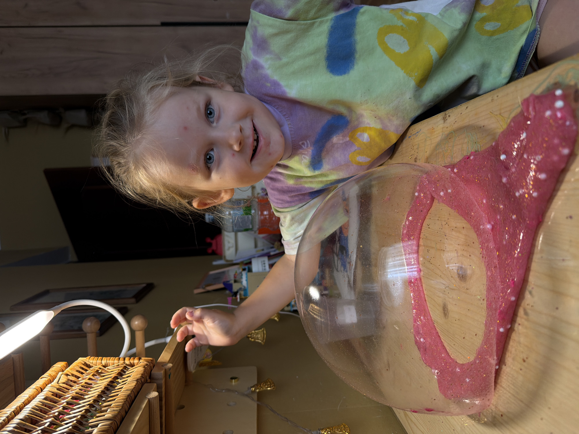 A young Caucasian girl with blonde hair and blue eyes, wearing a tie-dye t-shirt, is seated at a wooden table. She has red marks on her forehead and cheeks. In front of her on the table is a pink, glittery slime partially enclosed by a clear plastic dome. To her left, a wicker basket is attached to a shelf, illuminated by a bright LED lamp. Behind the girl, a wooden cabinet is visible. The scene appears to be indoors, possibly a home or classroom environment, in Khimki, Russia.