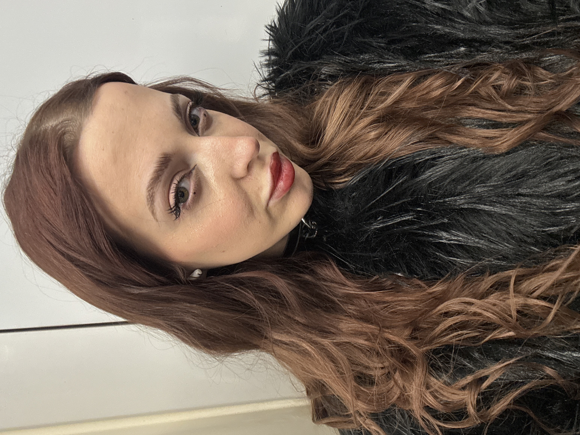 A woman with long, auburn-colored hair and fair skin is the subject of the image. She is wearing a black, furry coat or jacket. Her makeup includes lipstick and subtle eye makeup. She has light-colored eyes. The background appears to be a plain, white wall. There's no indication of Moscow, Russia. The scene is indoors.