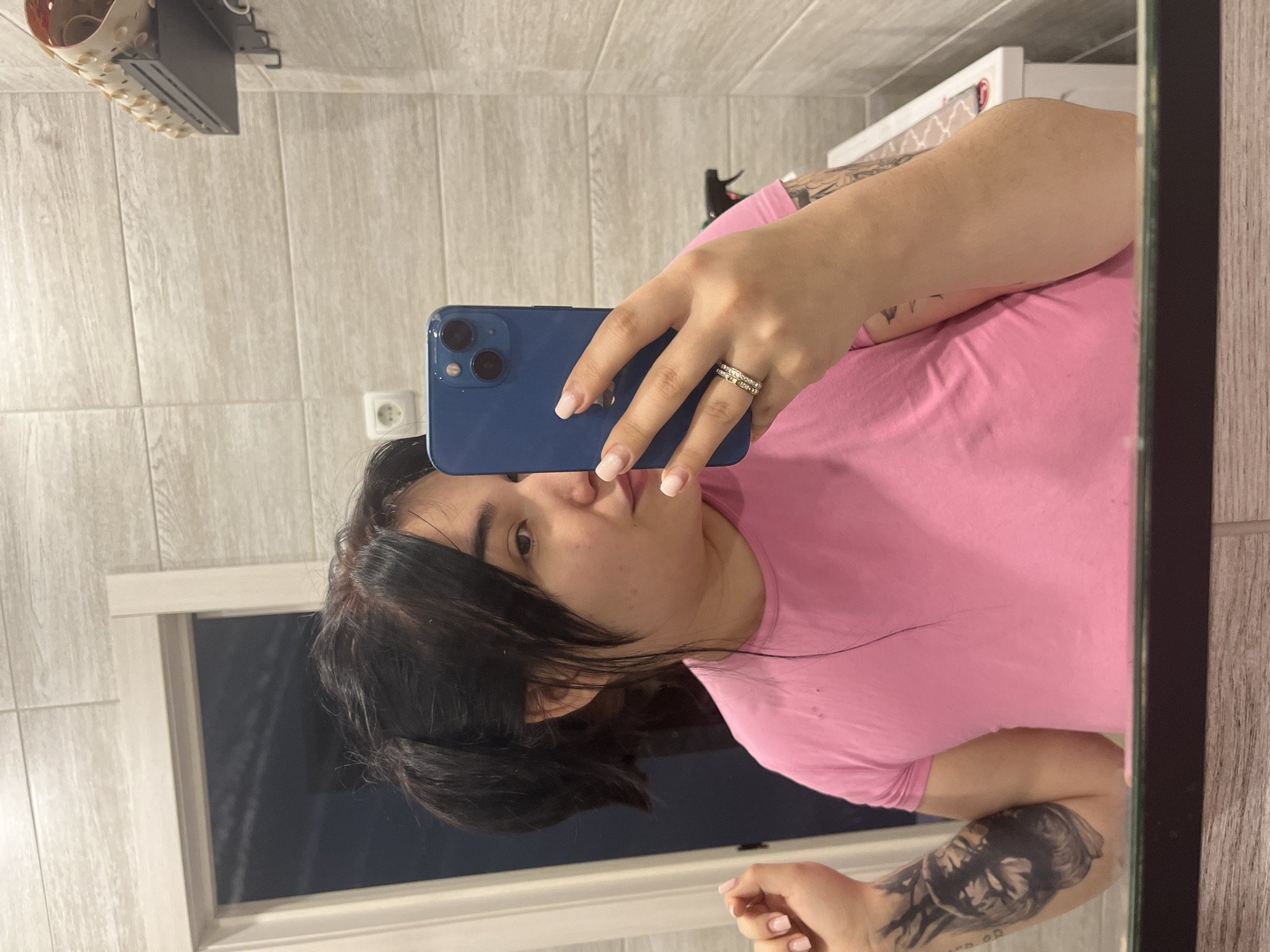 A person with dark hair is taking a selfie in a bathroom mirror. The person is wearing a pink top and holding a blue iPhone in their right hand. The bathroom has light wood-look tiled walls. A towel rack with towels is visible above. A window or dark surface is seen in the reflection behind the person. The person is wearing two rings on their ring finger and has pale pink nail polish. The person has a tattoo on their left arm. There is an electrical outlet on the wall. The location appears to be a domestic bathroom. There is no information to suggest this location is in Chaikovsky, Russia.