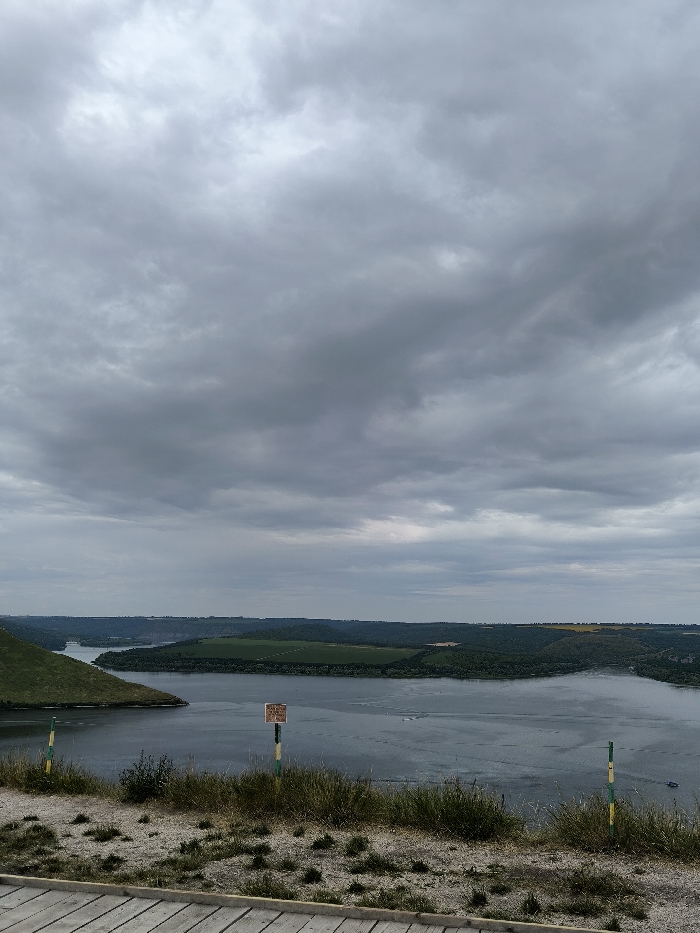 The image displays an elevated panoramic landscape view of the Dniester River canyon, located near the village of Horaivka, Khmelnytskyi Oblast, Ukraine. The scene is observed under a completely overcast sky filled with dense, uniform grey clouds.
In the immediate foreground, the bottom left shows a portion of a light-colored wooden plank surface, consistent with a boardwalk or viewing platform. The ground directly in front of the viewpoint is composed of light-grey, dry soil or gravel with scattered patches of green grass and weeds. Three slender poles are visible: one partially obscured on the far left with green and yellow stripes, a central weathered wooden post bearing a rectangular, light-brown sign with dark text, and another green and yellow striped pole on the far right, partially obscured by vegetation.
The midground is dominated by the wide, calm expanse of the Dniester River, which flows from the left of the frame and curves towards the right. The river's surface exhibits subtle ripples and what appear to be distant wakes from a small, unidentified watercraft, barely visible in the upper right quadrant of the water body. The river is bordered by steep, verdant hills and densely forested areas on the opposite bank.
The background extends into a series of undulating hills and plateaus, covered with a mosaic of dark green foliage and lighter green or yellowish agricultural fields. The distant horizon merges indistinctly with the low cloud base. No individuals or discernable structures other than the poles and implied platform are present in the immediate vicinity.
