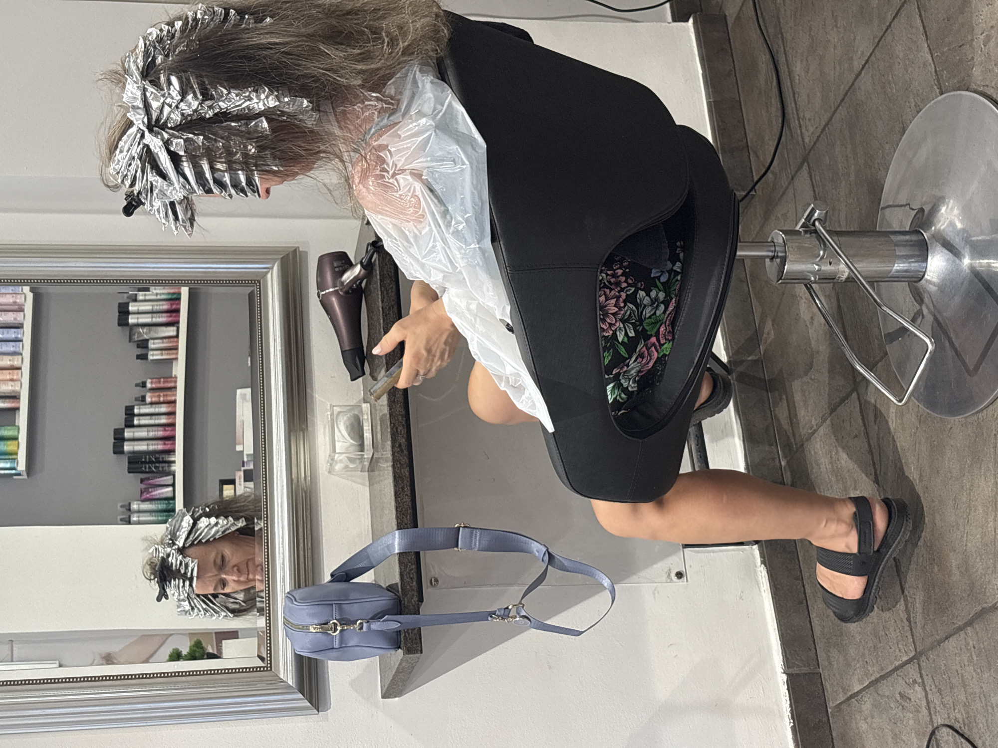 In Rome, Italy, a woman sits in a salon chair, her hair wrapped in aluminum foil for a coloring treatment. She is holding a smartphone and looking at it. A blue handbag hangs from a nearby shelf, and a hairdryer rests on the counter. The salon has white walls and a tiled floor. The woman is wearing a floral-patterned seat cover on her chair and black sandals. A large mirror reflects her face, also covered in foil.
