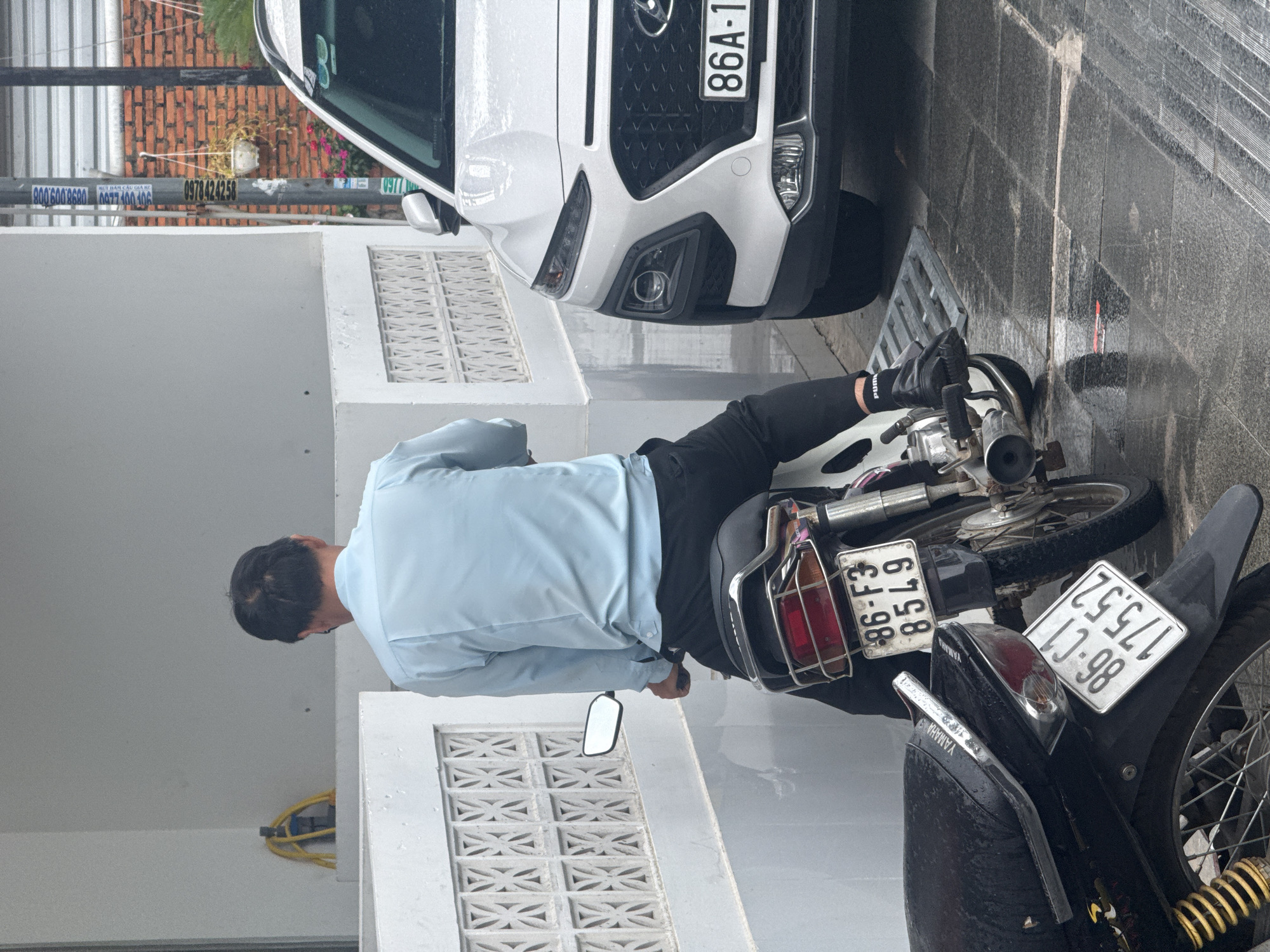 A man wearing a light blue long-sleeved shirt and black pants is seated on a motorcycle. The motorcycle has a visible license plate with the characters "86-F3 8549". Another motorcycle, predominantly black, is parked in front of the man's, with its license plate reading "86-C1 175.52". To the right of the motorcycles is a white car, possibly a Hyundai Kona, with a license plate displaying "86A-1". The scene appears to be outdoors, with a wet, tiled ground suggesting recent rain. A white decorative wall or railing is visible to the left, and a brick wall with a hanging potted plant is in the background to the right. A yellow hose is coiled on a ledge in the far left background. The overall setting is an urban street in Phu Thuy Ward, Vietnam.
