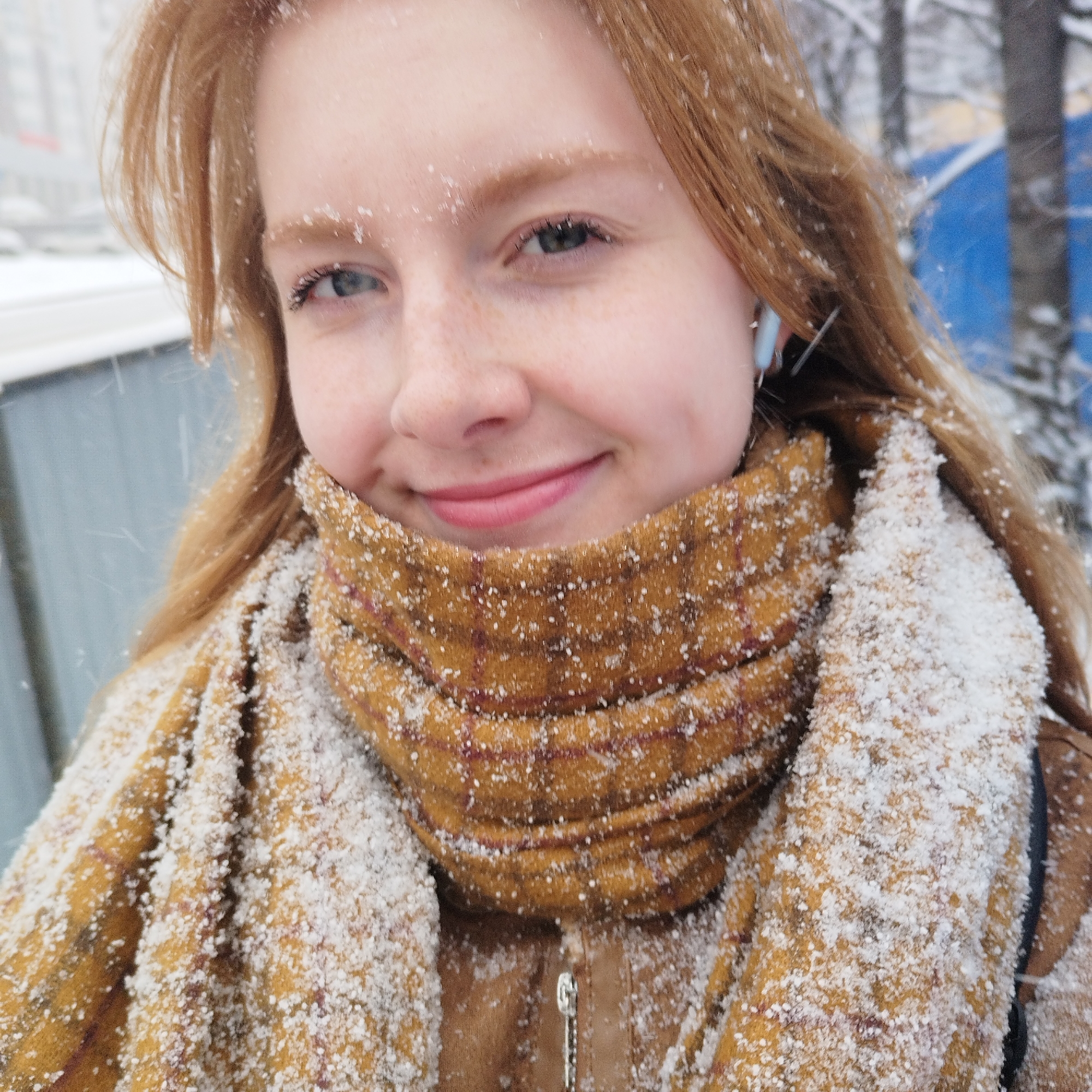 A young woman with red hair is smiling at the camera in a snowy environment. She is wearing a brown jacket and a brown and yellow plaid scarf. Snowflakes are visible on her hair, face, and scarf. In the background, there are buildings and trees covered in snow. The image was taken in Yekaterinburg, Russia.