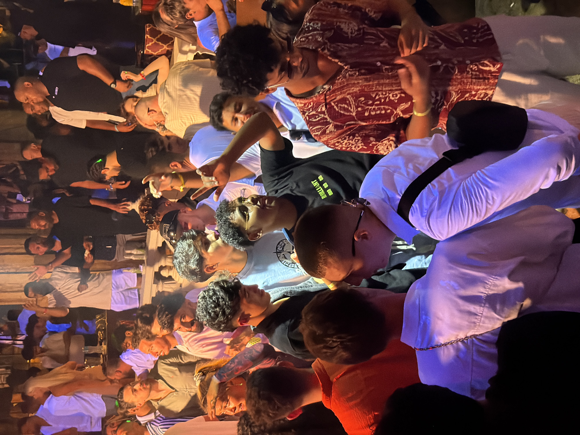 The image depicts a crowded outdoor event, likely a nightclub or beach party, illuminated by artificial lighting in Sharm El Sheikh, Egypt. A diverse group of young adults are visible, many dressed in casual attire such as t-shirts, shirts, and shorts. Several individuals are engaged in conversations and social interactions, with some appearing to be dancing or moving within the throng. The environment suggests a lively and energetic atmosphere. The background reveals architectural elements and possibly stage lighting, indicative of a commercial entertainment venue. The overall scene captures a moment of collective enjoyment and social gathering in a popular tourist destination.