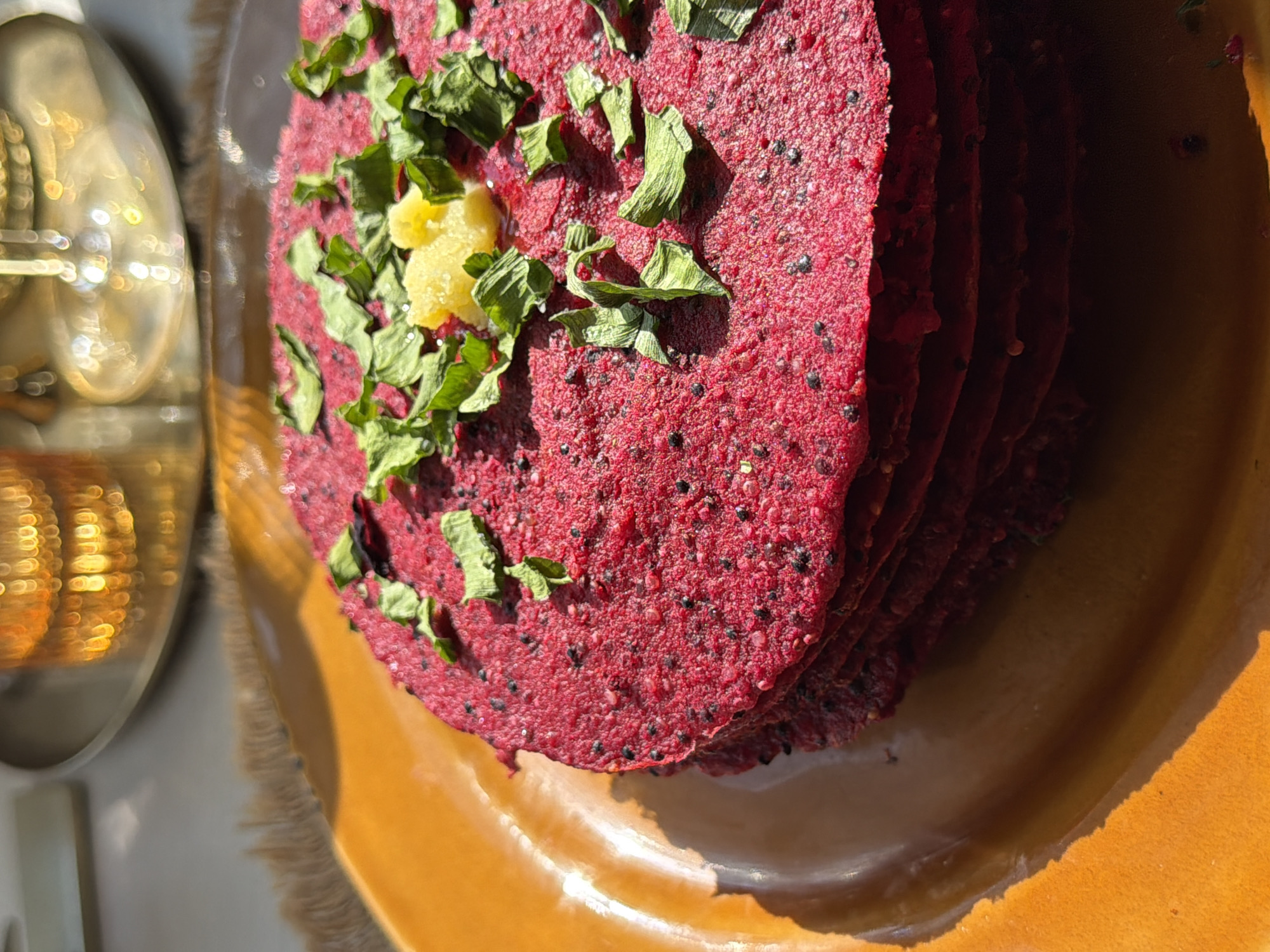 The image shows a stack of what appears to be beet pancakes on an orange plate. The pancakes are deep red with small black dots and topped with chopped green herbs and a small dollop of yellow butter or cheese. The plate sits in sunlight, creating a strong shadow. To the left, there is a blurry reflection of a golden object, possibly silverware or a decorative piece. No people are visible in the image. The location in the city of Krasnoyarsk, Russia, is not discernible from the image.