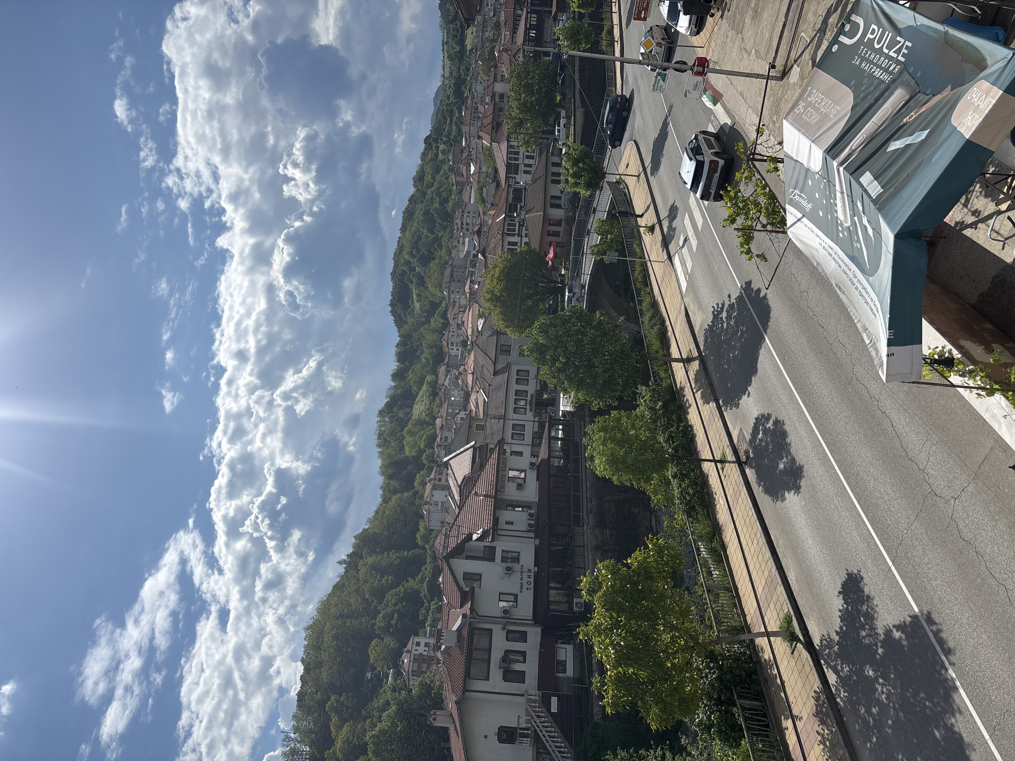 The image shows a view from above in Zlatograd, Bulgaria. A hillside densely covered with houses and trees rises to the left. The sky is blue with dramatic, puffy white clouds. To the right is a road with sparse traffic, and a sidewalk. A "PULZE" branded structure stands on the right. Buildings, including one identified as "ТОНИ КЪЩА ЗА ГОСТИ," dot the hillside.