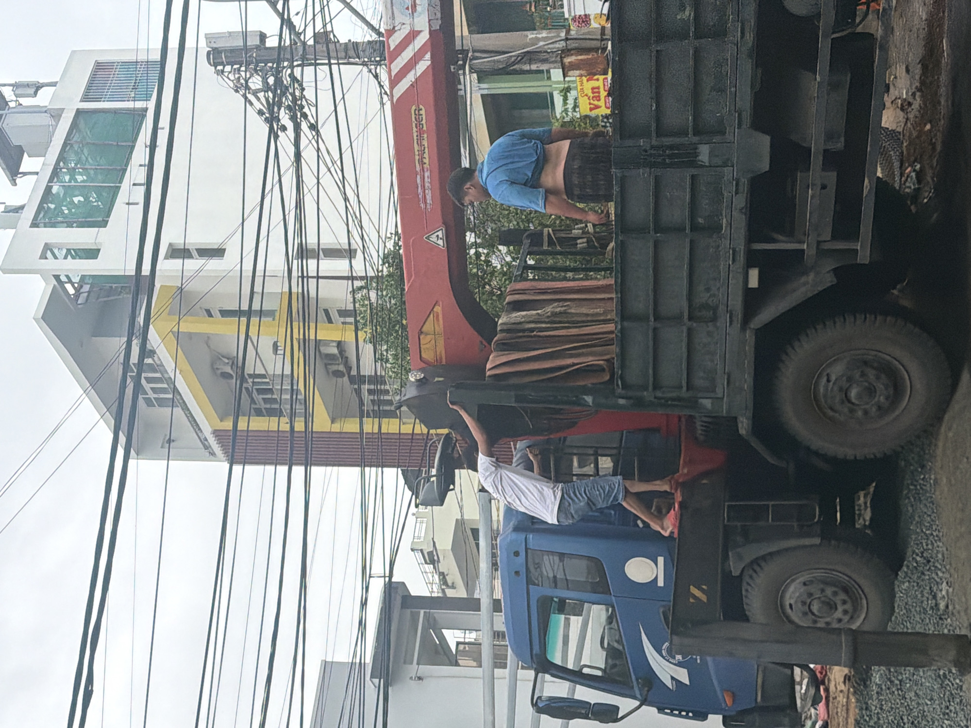 This daytime image, set in Nha Trang Ward, Vietnam, captures an urban street scene centered around a blue flatbed truck equipped with a red hydraulic crane arm. The truck is parked on gravelly or unpaved ground, and two men are actively engaged with it. One man, wearing a blue t-shirt and dark shorts, stands in the truck's bed, appearing to handle materials or operate near the base of the crane. The second man, in a white t-shirt and dark shorts, is positioned on the side step of the truck's cabin, holding onto the vehicle, likely assisting or maneuvering.
In the background, under an overcast sky, are multi-story buildings, including a prominent white structure with green-tinted windows and another building with yellow trim. A dense network of black overhead power lines and cables crisscrosses the upper part of the frame, strung between utility poles and buildings, characteristic of many Vietnamese urban areas.
Notable visible text includes "KATO" and a warning triangle symbol on the red crane arm. A partially visible yellow sign in the background displays Vietnamese text, reading "CUA HANG" (Shop) and "Vân N". The scene suggests a workday activity involving the truck and its crane, possibly for loading or unloading.
