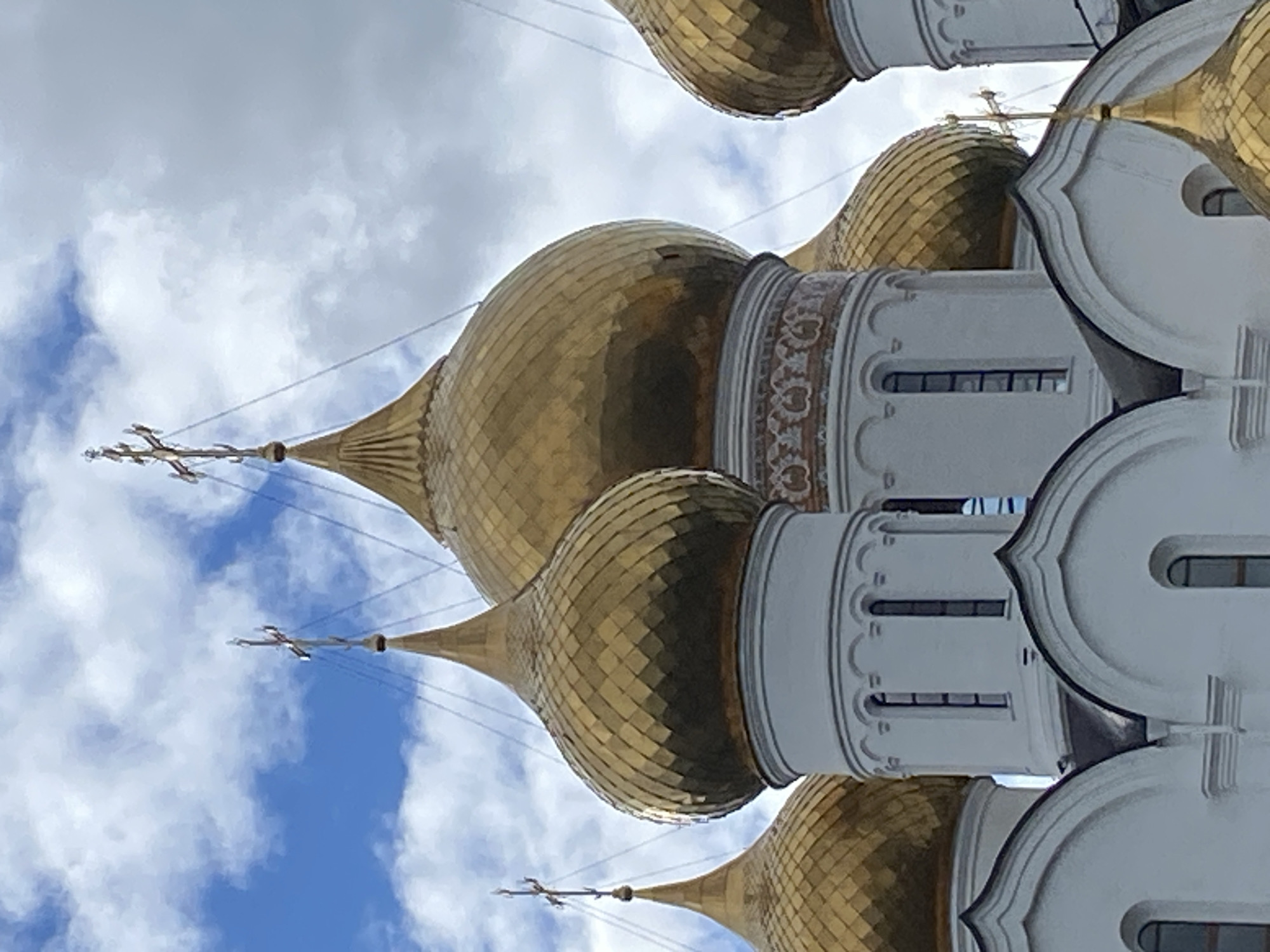 The image shows a close-up view of a church with multiple golden domes against a backdrop of blue sky and clouds. The church is characterized by its white walls and decorative elements. The domes are covered in gold tiles. Thin power lines are visible in the background, partially obscuring the view of the sky and domes.
The location is in Заволжье, Russia.