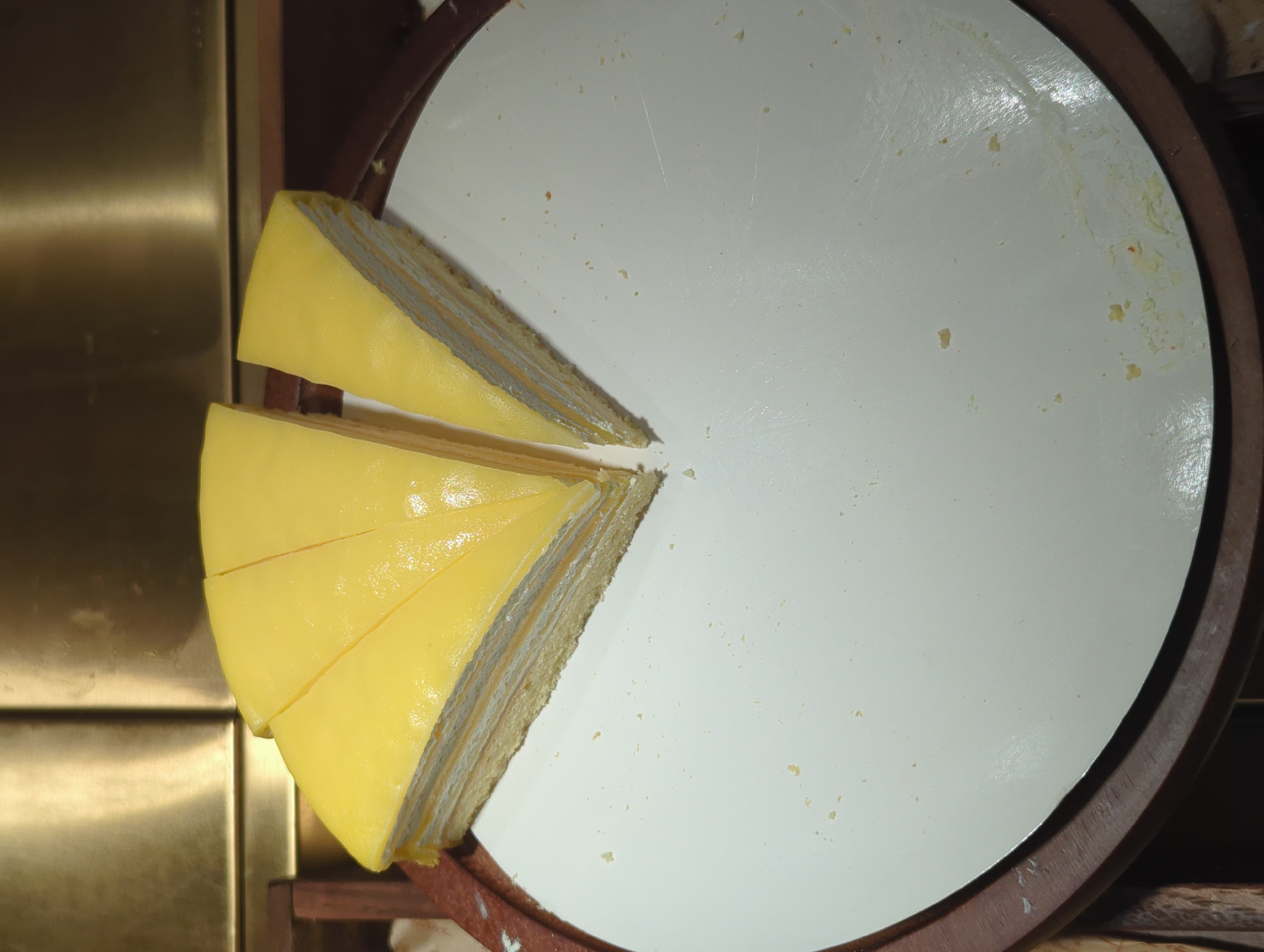 A sliced cake with a yellow frosting and layers of cream and cake is displayed on a white plate, which sits on a dark wooden base. The cake is cut into wedges, with some pieces missing, leaving crumbs on the plate. The background features a metallic, textured surface. The setting appears to be indoors, possibly a bakery or a restaurant in Yuzhong District, China. There are no people visible in the image.