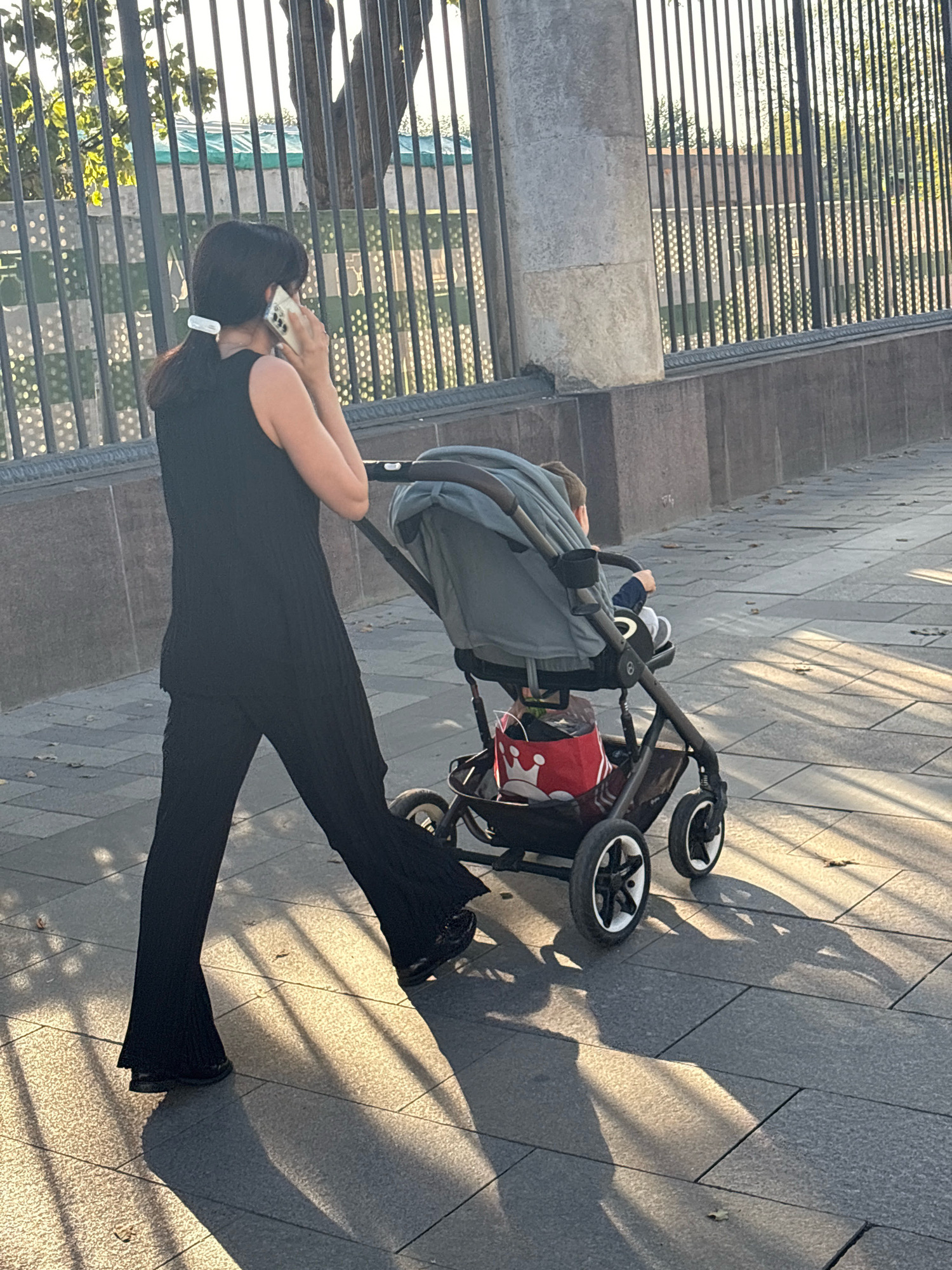 A person is pushing a stroller with a child in it on a sidewalk in Moscow, Russia. The person is wearing a black sleeveless top and black flared pants. They are holding a mobile phone to their ear and appear to be in conversation. The stroller is gray and has a red bag with a white crown design in the bottom basket. The child is wearing a blue jacket and a white hat. The sidewalk is made of gray stone tiles, and there are shadows cast by a fence and trees. A metal fence is visible in the background. The sun is shining, creating long shadows on the ground.