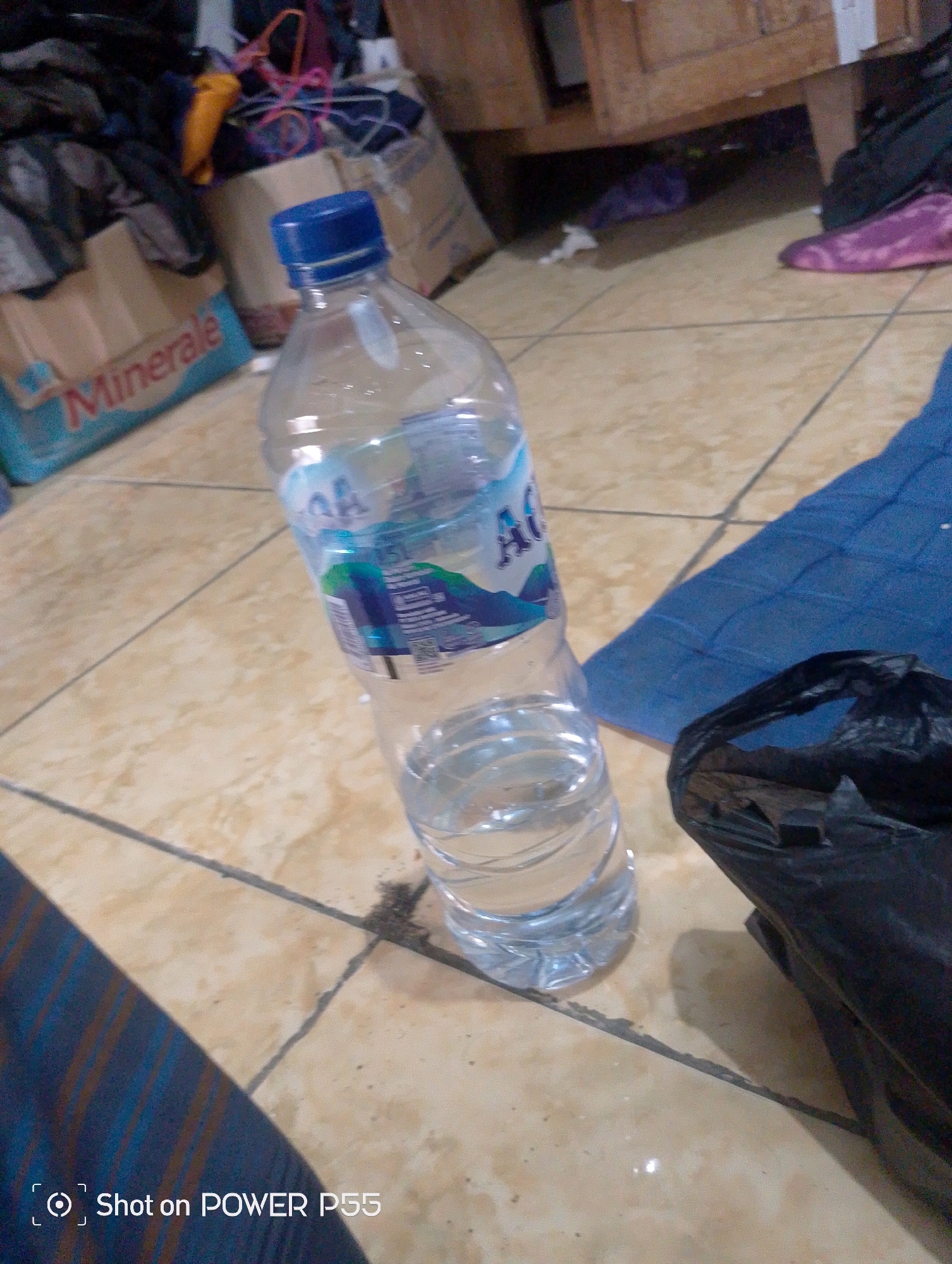 A partially full 1.5-liter bottle of "Aqua" mineral water stands upright on a tiled floor. The bottle has a blue cap and a label with blue and green graphics. To the left of the bottle, a cardboard box labeled "Minerale" is visible. Behind the bottle, a pile of clothing and other items are scattered, and a dark blue cloth is spread on the floor. A black plastic bag is situated to the right of the water bottle. The lighting suggests it is indoors during the day. The image contains a watermark in the bottom left corner: "Shot on POWER P55". The scene is likely a domestic setting within the city of Lebaksiu, Indonesia, given the presence of Indonesian brands and the watermark.