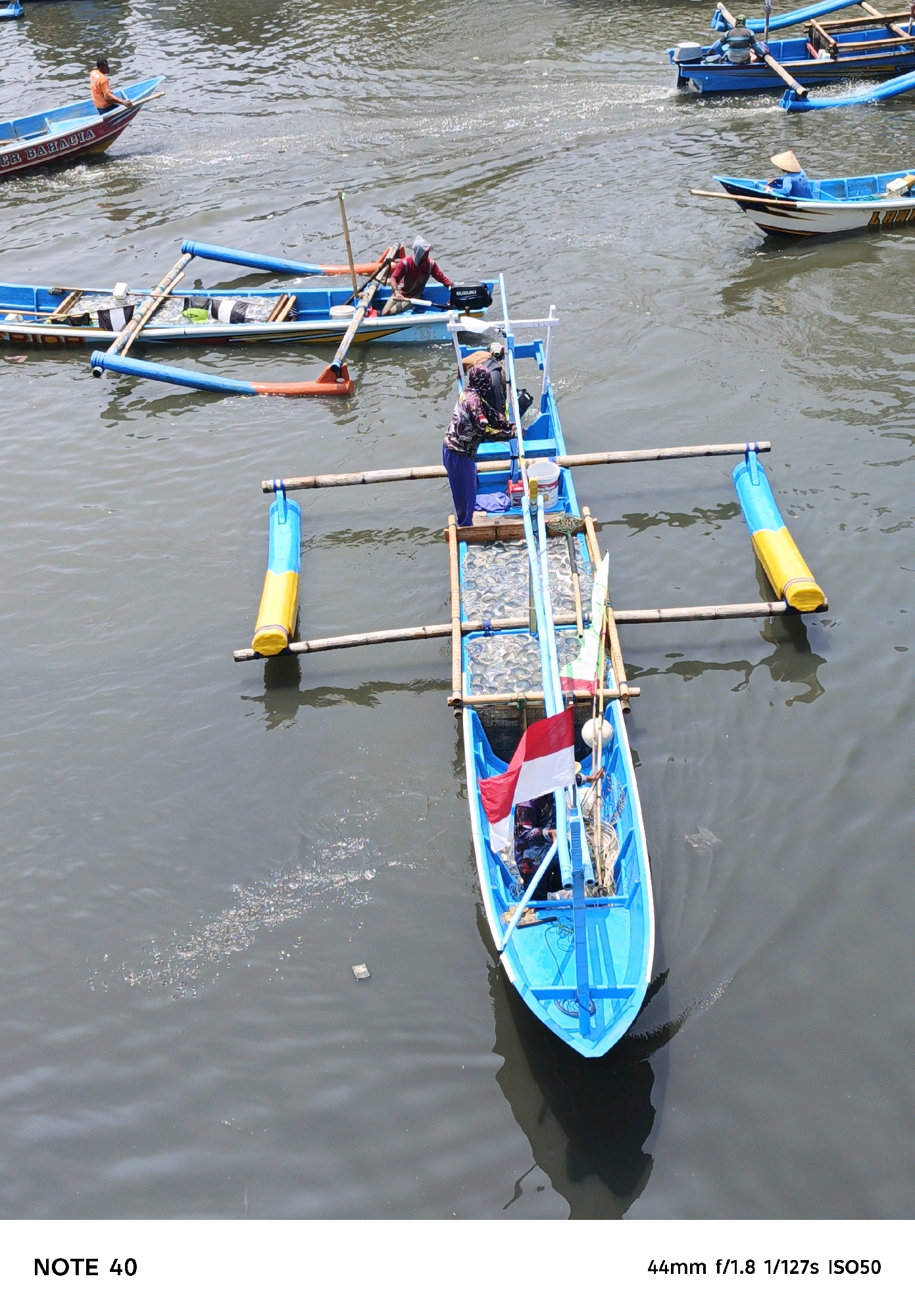 An overhead shot captures several small blue and white boats in the murky waters of Kelurahan Tegalkamulyan, Cilacap, Indonesia. In the foreground, a bright blue boat with a red and white Indonesian flag is prominent. It is equipped with bamboo outriggers and carries a person wearing a dark shirt and blue pants. The boat appears to be transporting a catch of numerous small fish in two rectangular containers. A person in a red shirt and head covering works on another boat nearby, which also has bamboo outriggers and appears to be a similar type of fishing vessel. Further in the background, two more boats are visible. One is a larger blue boat with a person in an orange shirt on board, and the other is a smaller blue and white boat with a person wearing a hat. The water is filled with debris, indicating a potentially polluted environment.