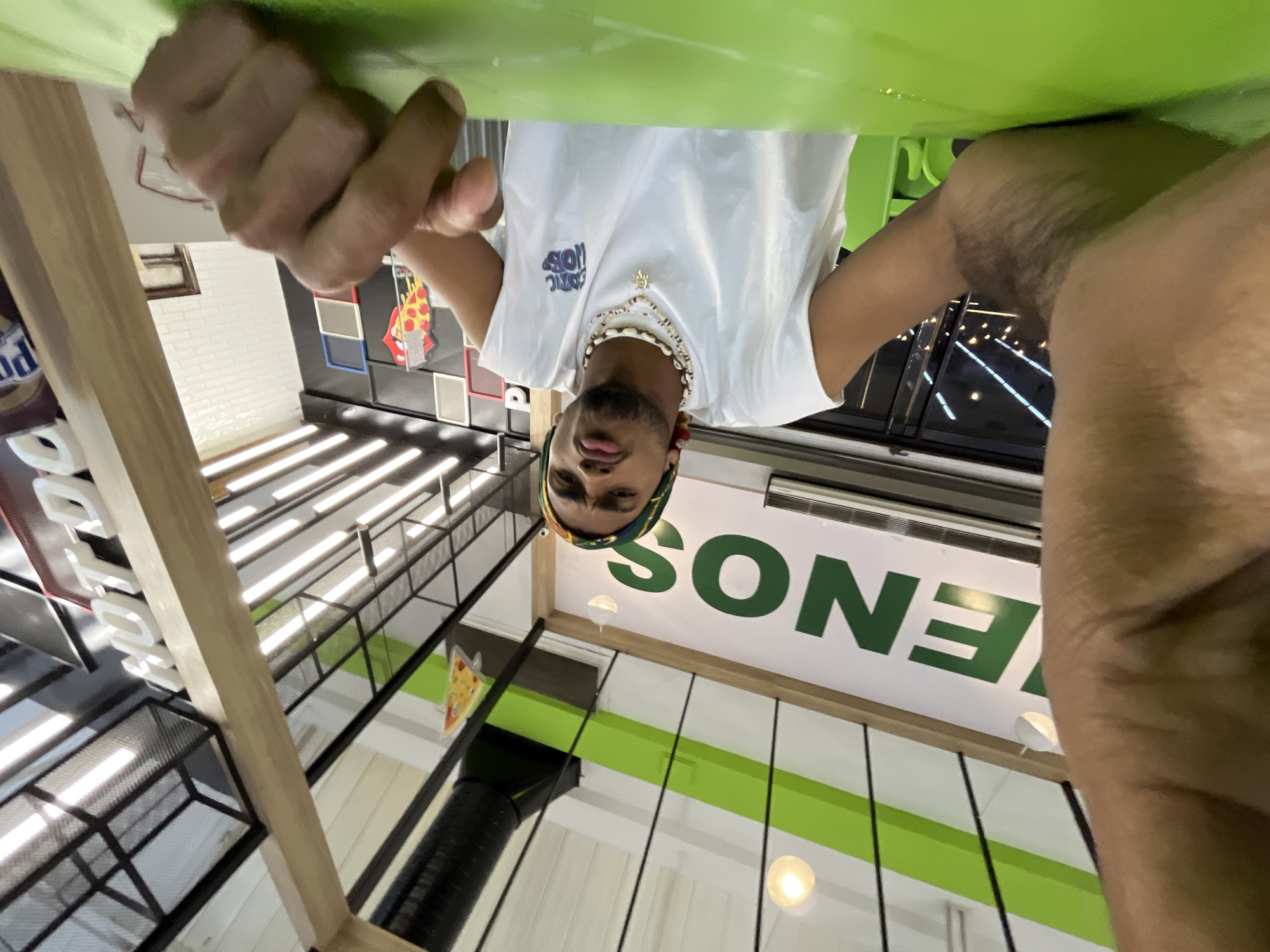 A man wearing a white t-shirt, beaded necklaces, and a head wrap is upside down in a brightly lit interior space. He is holding onto a green surface above him, obscuring the top of the image with his hands and arms. In the background, the word "SONES" is partially visible on a white wall. The setting appears to be a commercial establishment with modern design elements, including exposed ductwork, metal framework, and decorative pizza-themed wall art. There is no explicit information to confirm the location is in Porlamar, Venezuela.