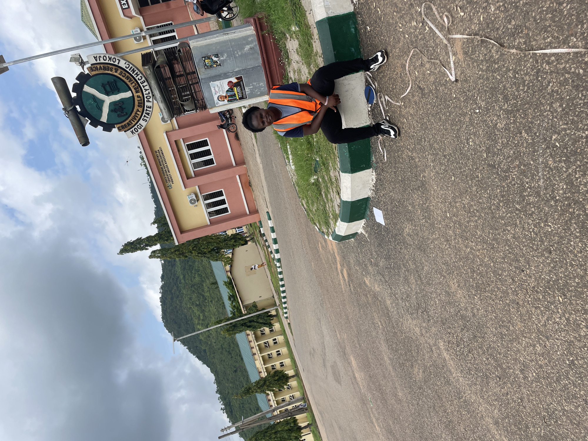 A young woman wearing an orange safety vest over a blue shirt and black pants sits on a curb outside a building at Kogi State Polytechnic, Lokoja, Nigeria. The woman is looking at the camera with a slight smile. She is wearing black and white sneakers. A tape measure is spread out on the asphalt in front of her. In the background, a building with pink and yellow accents is visible, along with a large sign for Kogi State Polytechnic. A lush green mountain is visible behind the buildings. The sky is partly cloudy. The scene appears to be outdoors on a paved area.