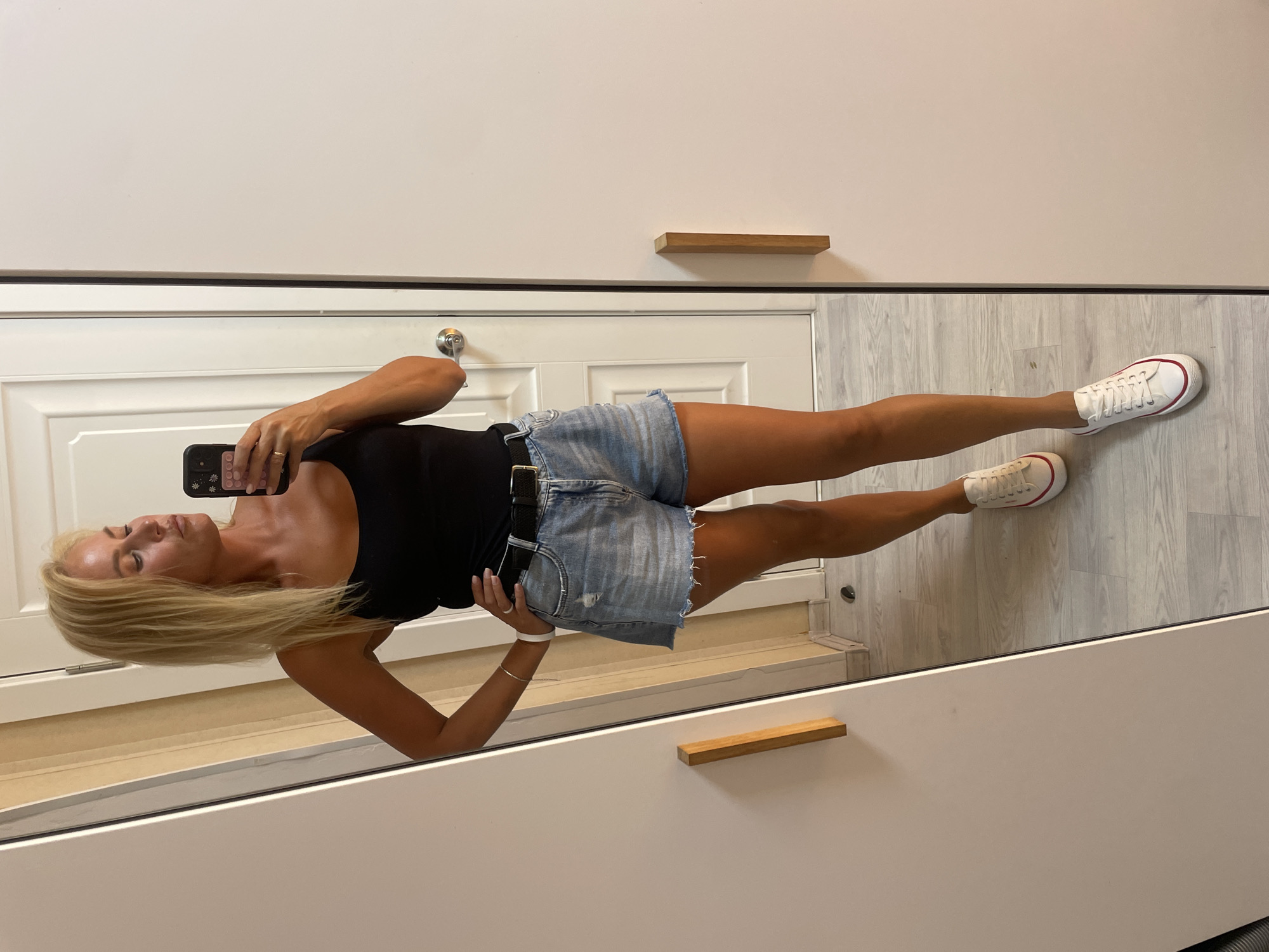 A blonde woman is taking a mirror selfie indoors. She is wearing a black tank top, denim shorts, a black belt, and white sneakers with red trim. She has her left hand on her hip and is holding a smartphone in her right hand. The background shows a closet with wooden handles, a door, and a wooden floor. The location is not specified in the image.