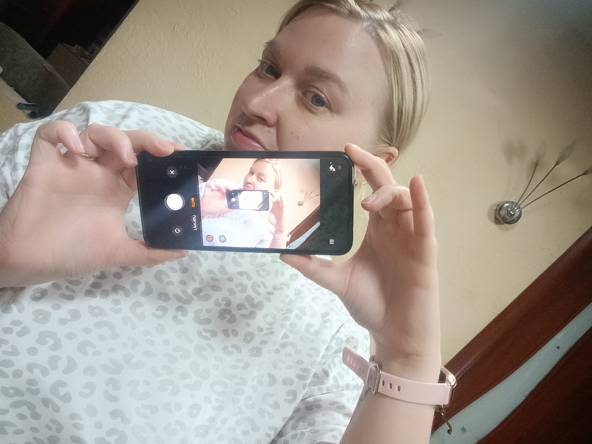 A blonde woman with blue eyes takes a selfie in an indoor setting. She holds a black smartphone horizontally, with its screen displaying a recursive image of herself taking the picture, creating an "infinity mirror" effect. Her expression is neutral as she looks towards the phone's camera.
She is wearing a light-colored top with a subtle leopard print. A gold ring is visible on her left hand's index finger, which is holding the phone. On her right wrist, a pink smartwatch with a gold-colored clasp is visible.
The phone's camera interface is active, showing a large white shutter button, an 'X' icon for closing, and text labels in Cyrillic: "ФОТО" (PHOTO) and "ПОРТРЕТ" (PORTRAIT), indicating camera modes.
The background consists of a plain light-colored wall. To the left, dark furniture, possibly a couch or bed, is partially visible. To the right, a distinctive decorative object resembling a clock or art piece with multiple slender arms protrudes from the wall. The lighting appears soft, typical of an indoor environment, possibly daytime with ambient light or artificial illumination.
