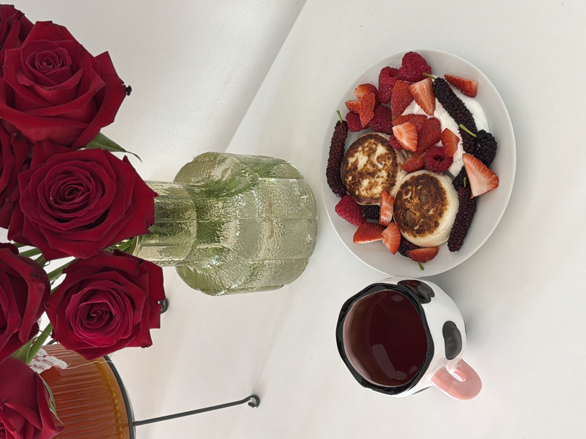 The image is a flat lay composition depicting a breakfast or brunch setting.
On a white surface, likely a table, are several items:
1. **Roses:** A bouquet of deep red roses in a vase. The vase appears to be made of patterned glass.
2. **Food:** A white plate contains what appears to be small pancakes or syrniki, adorned with fresh raspberries, sliced strawberries, dark mulberries, and a dollop of white cream.
3. **Drink:** A black-and-white cow-patterned mug is filled with a dark liquid, possibly coffee or tea. The mug features a pink handle.
4. **Background:** The wall is white and has a minimalist aesthetic.
Given the type of food (syrniki) and the style of the image, it is plausible that the scene is from a location, possibly a home or café, in Tel Aviv, Israel, where international and local culinary trends are common.