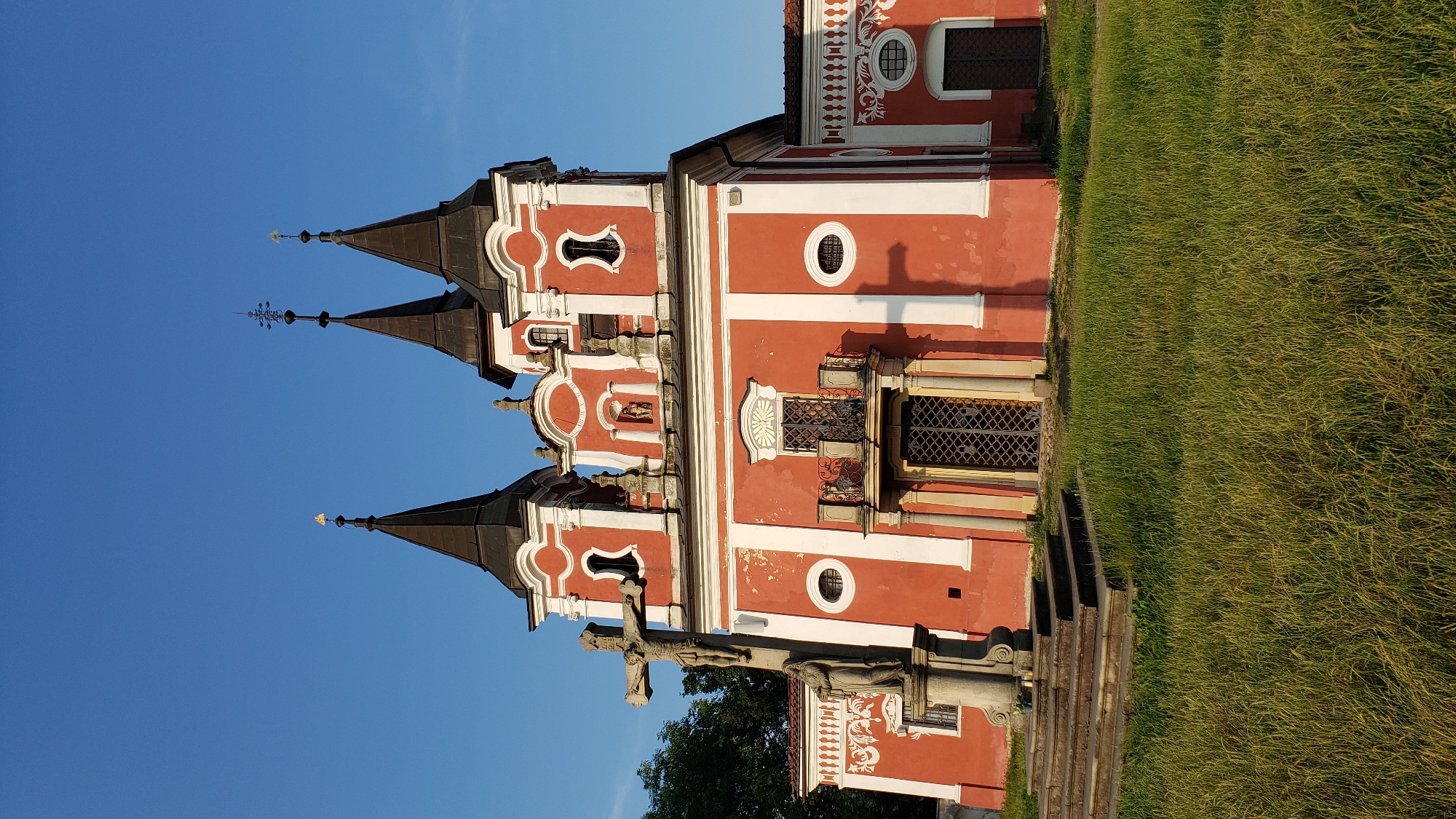 The image depicts the Church of Saint Nicholas in Prešov, Slovakia, under a clear blue sky. The church is painted a vibrant red with white accents and features three prominent towers, each topped with a dark spire. In front of the church stands a stone crucifixion statue with Christ on the cross and a figure of a woman at the base. A set of stone stairs leads up to the church entrance, which is a double door with ornate metalwork. The area surrounding the church is covered in lush green grass. The overall scene suggests a peaceful and well-maintained religious site.