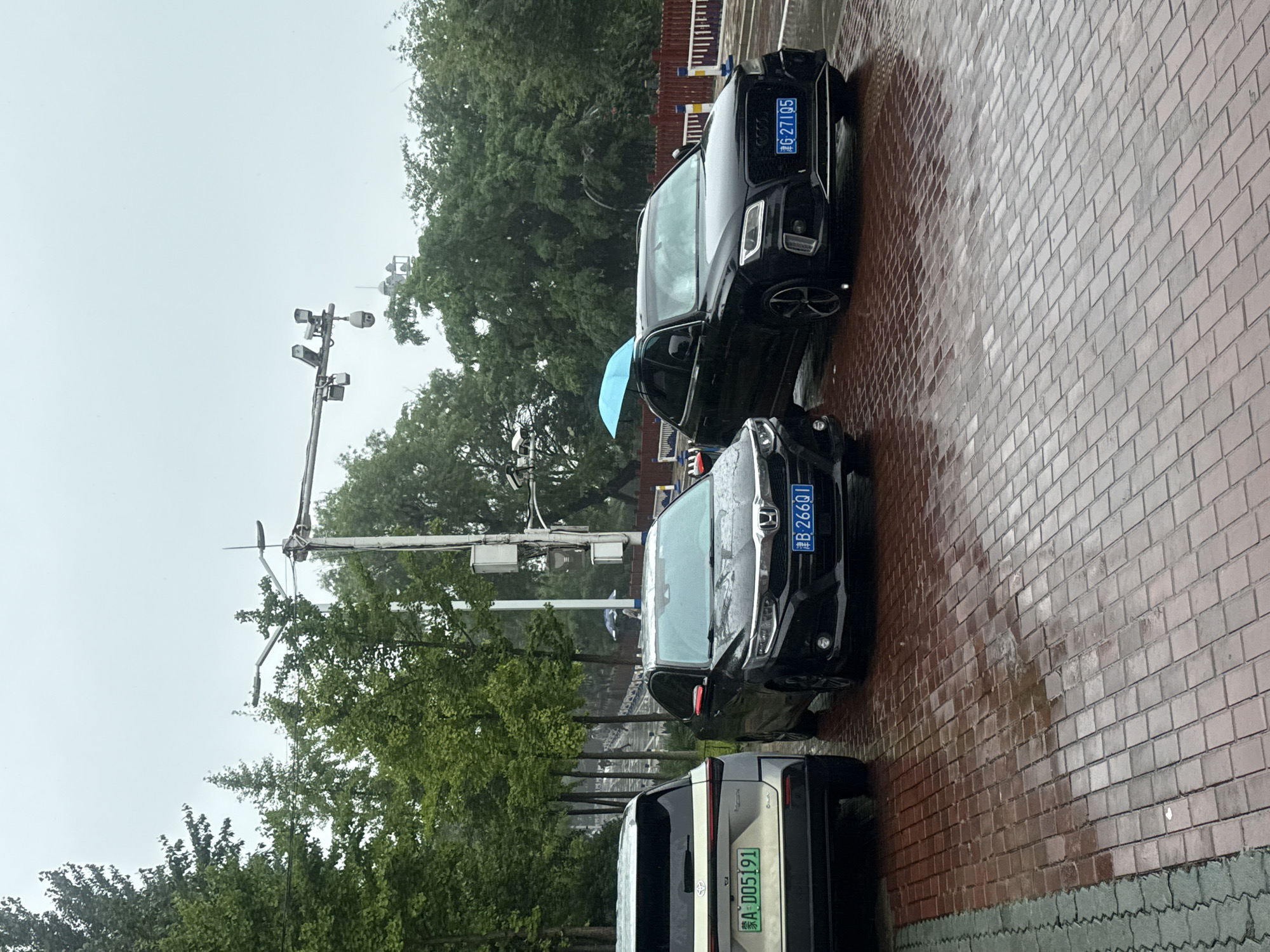 In Qinhuangdao, China, on a cloudy and rainy day, three cars are parked on a wet, red brick-paved area. The car on the left is a silver SUV with a license plate reading "A D05191". In the center, a black sedan with a license plate "B 266Q1" is parked. To the right, a black SUV has a light blue umbrella open on its roof and a license plate reading "G 271Q5". Behind the cars, trees line a path with a bridge visible in the distance. Traffic lights and security cameras are mounted on poles overhead.