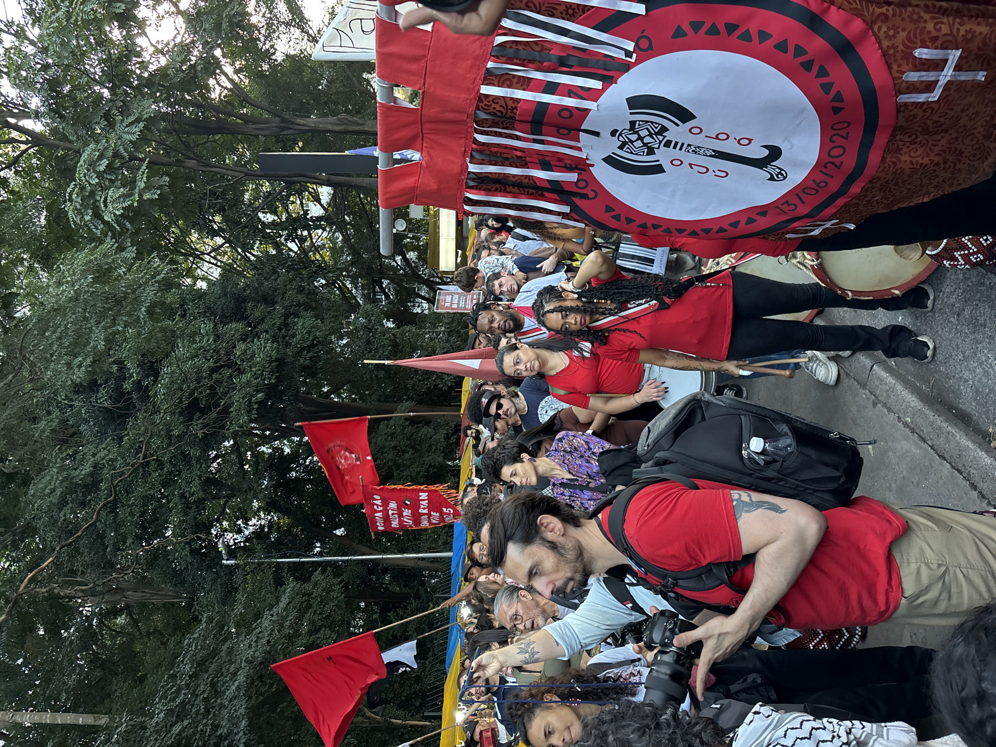 A street-level, medium-shot depicts a demonstration or protest in what appears to be the city of São Paulo, Brazil. The scene unfolds vertically, with the foreground closest to the viewer and the background receding into the distance. A crowd of people occupies the lower portion of the frame, including a man in the front with a red shirt and a black camera, suggesting he may be a photographer. Red flags are interspersed among the crowd, along with a banner featuring the date "13/06/2020" and what appears to be tribal symbols. The participants are engaged in a march or demonstration, with varying degrees of visible emotion. The trees lining the street give the area a verdant appearance, typical of urban parks or tree-lined avenues. The natural light suggests it is daytime, adding to the scene's vibrancy. The elements combined indicate an organized political or social movement expressing solidarity or raising awareness in an urban setting.