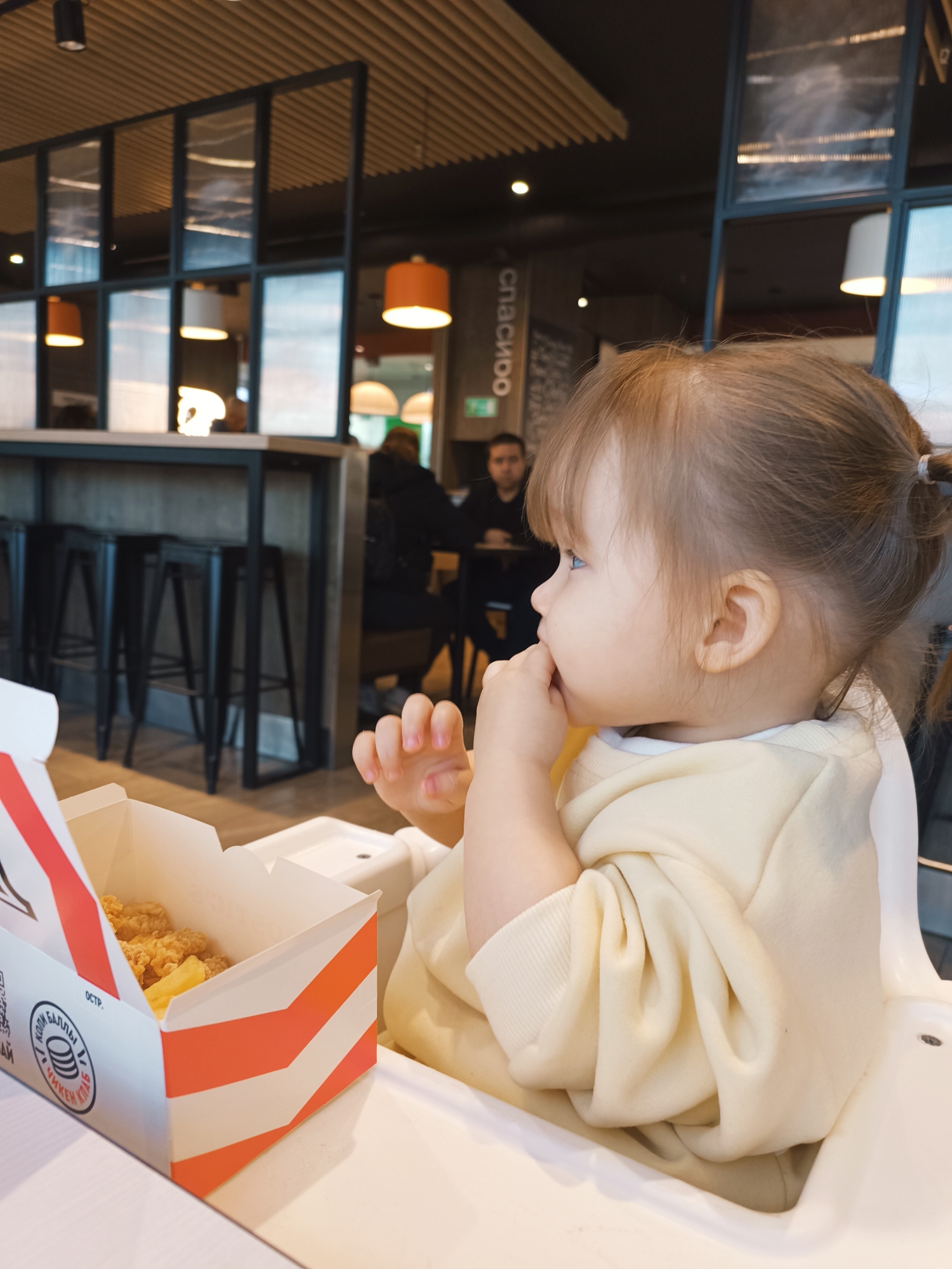 A young child is seated in a white highchair at a table in a restaurant in Izhevsk, Russia. The child, with light skin, brown hair in a ponytail, and wearing a light yellow sweatshirt, is looking to the left with fingers near the mouth. A white and orange striped box containing fried food is on the table to the left of the child. In the background, there are black bar stools, a partially visible man in a black shirt, and window partitions with reflective glass. A sign with Cyrillic script is also visible. Overhead, the restaurant has a slatted ceiling and hanging light fixtures.