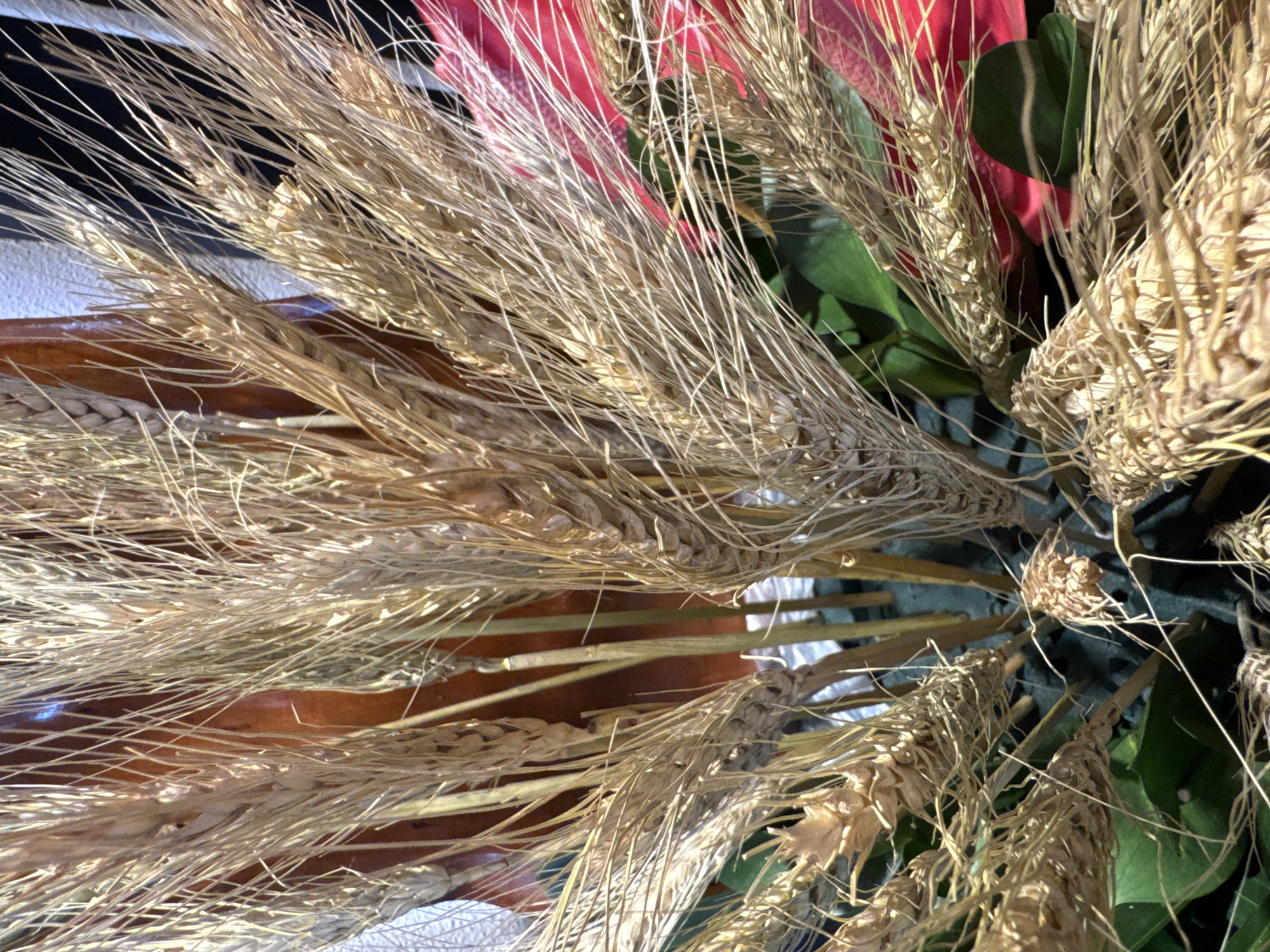 The image depicts a close-up, overhead shot of a floral arrangement or bouquet. The arrangement consists primarily of dried wheat stalks, with some green leaves and hints of red flowers visible amongst them. The stalks are a light golden-brown color, and the overall composition is dense and slightly cluttered. There is a reflective surface beneath some of the stalks, possibly a tabletop. The setting appears to be indoors, and based on the prompt constraints, it is presumably in Kuta Selatan, Indonesia.