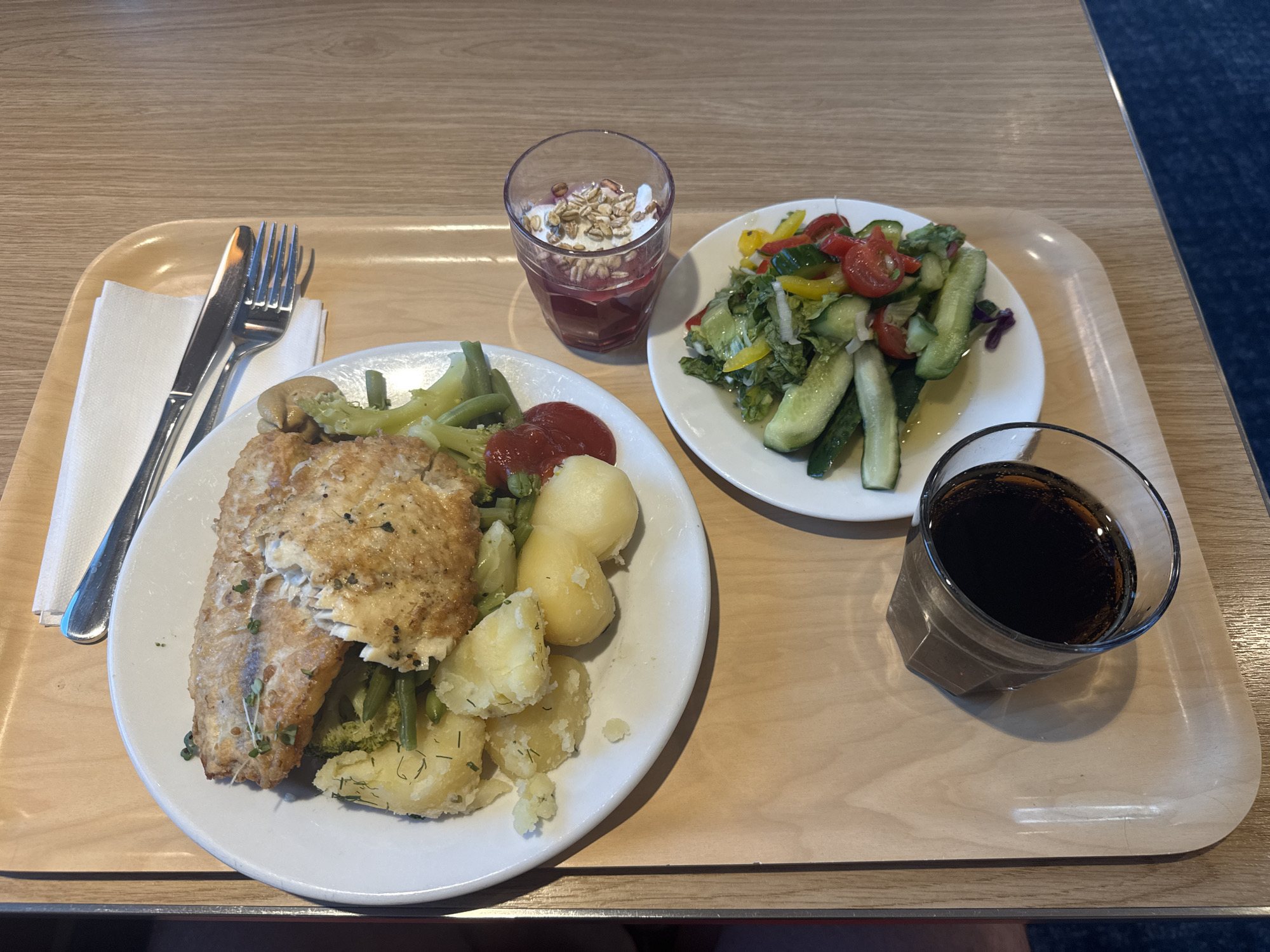 The image shows a wooden tray on a table, laid out with a meal and a drink. On the tray, there is a white plate with a portion of pan-fried fish, served with boiled potatoes, broccoli, and green beans. Next to this plate is a small glass containing a layered dessert, possibly yogurt with granola and a fruit compote. There is also a side plate with a salad composed of cucumbers, tomatoes, lettuce, and peppers. A glass of dark liquid, likely soda, is placed on the tray to the left of the salad. A fork and knife are positioned on top of a white napkin at the top of the tray. The meal appears to be set in a cafeteria or restaurant setting, given the presentation and the type of food. The lighting suggests an indoor environment. The question about the location in Ösmo, Sweden, cannot be answered from the visual information provided in the image.