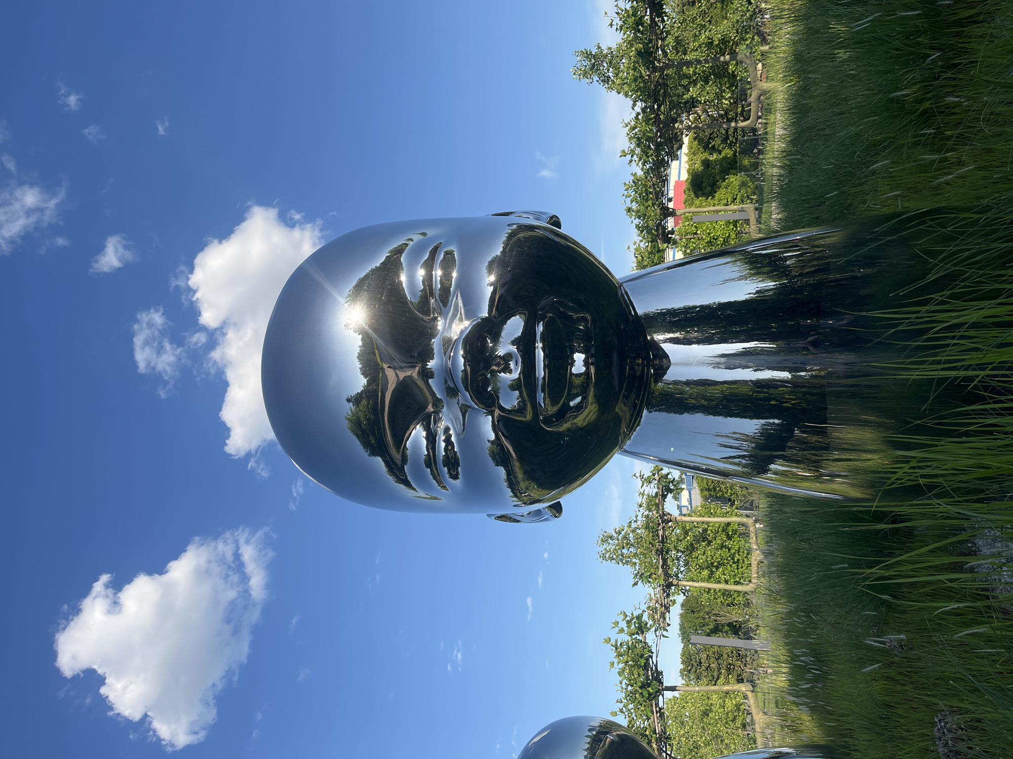 A large, highly reflective, chrome or stainless steel sculpture of a human head and the top of the shoulders is the central subject of this outdoor photograph, set in Krasnodar, Russia. The sculpture's mirror-like surface distorts and reflects its surroundings, including a vivid blue sky dotted with scattered white cumulus clouds. A bright sun glint creates a starburst effect on the sculpture's forehead, indicating strong midday or early afternoon light.
The background features lush green grass and mature trees, some of which appear to be espaliered or shaped. These elements are also visible as warped reflections on the sculpture itself, particularly on the contours of the face and cranium. The overall setting suggests a park or an outdoor art installation. No specific activity or event is taking place, and no people are visible in the immediate vicinity of the sculpture. The weather is clear and sunny. There is no visible text in the image.
