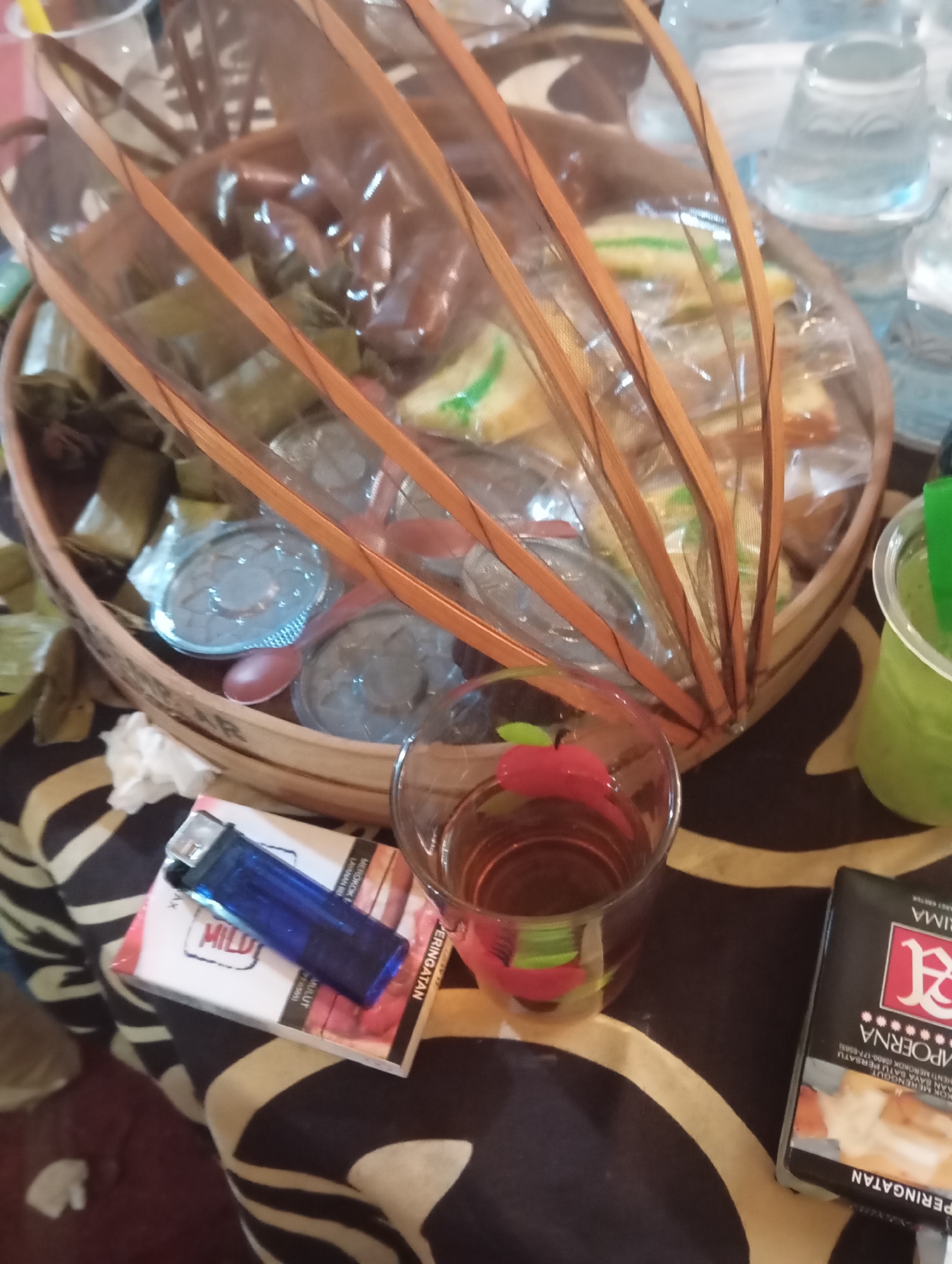 The image depicts a close-up, overhead view of a table setting. In the center, a woven basket contains individually wrapped snacks, possibly traditional Indonesian food items like lemper or arem-arem, covered by a clear plastic film supported by thin wooden slats. To the left, a blue lighter rests on a pack of cigarettes, which appears to be "MILD" brand. In the foreground, a glass filled with a dark amber liquid, likely tea, is visible, with a small red, apple-shaped charm hanging from its rim. To the right, a green beverage in a plastic cup with a straw is partially visible. The table is covered with a dark fabric featuring a repetitive pattern of alternating black and tan wavy stripes. In the background, several clear plastic cups are stacked. The scene suggests a casual gathering or a moment of respite. The location is identified as Kemangkon, Indonesia.