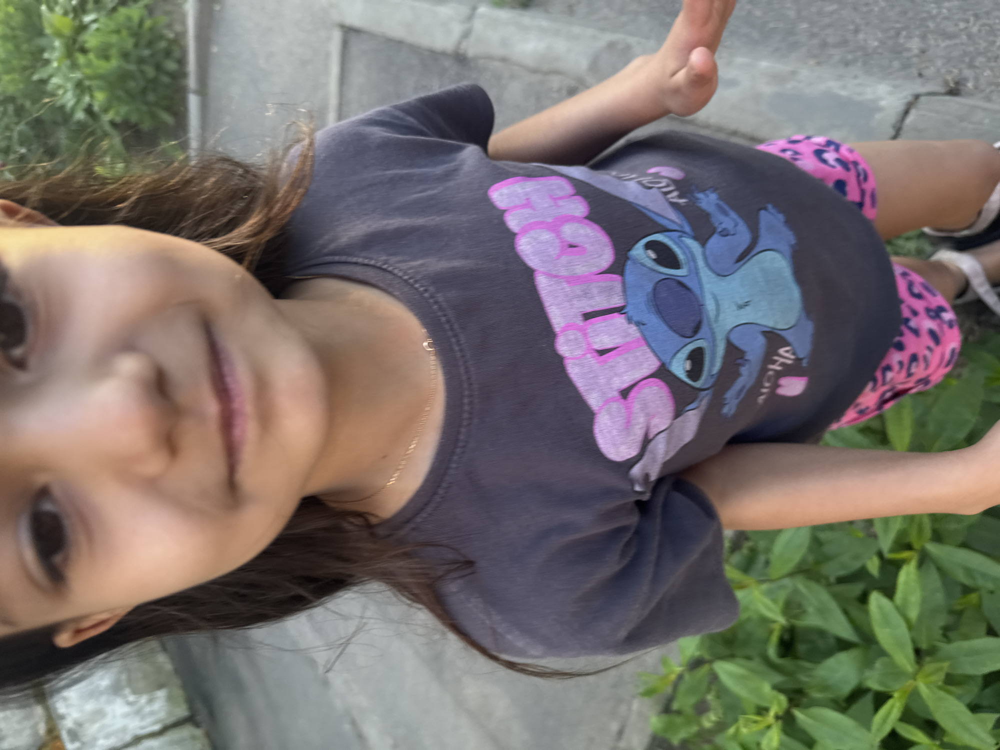 A young girl, viewed from the chest up, smiles at the camera. She has dark hair and a light complexion. She is wearing a dark gray t-shirt with a graphic of the cartoon character Stitch, and pink leopard-print shorts. The girl is standing outside, with green foliage to her left and a gray paved area to her right. The background is slightly blurred, suggesting movement or a shallow depth of field. The image appears to be taken outdoors during the day. The location is identified as Unknown, Unknown.