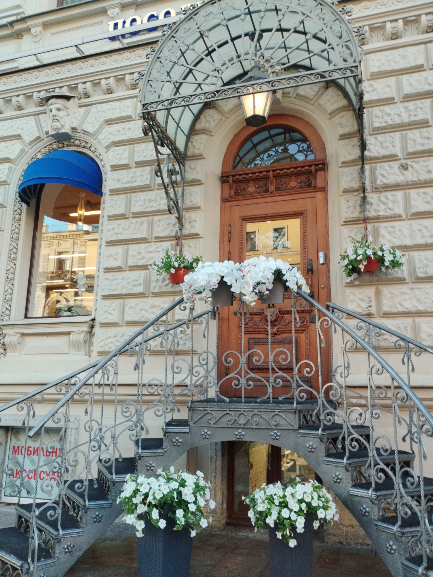 This is a medium shot, eye-level, exterior photograph of an ornate building entrance in Saint Petersburg, Russia. The building's facade is made of light-colored stone blocks, with decorative carvings around the arched doorway and window. A blue awning shades a window on the left, and a sculpture of a classical head is mounted above it. A wrought-iron canopy with a glass roof shelters the wooden double doors, and an old-fashioned lantern hangs from the canopy.
A dark metal staircase with decorative scrollwork leads up to the entrance. Two large planters filled with white petunias are placed on the steps. Another planter with white flowers hangs from a bracket on the wall to the right of the door, and a similar planter is visible to the left of the door. The building's reflection is visible in the window. Below the stairs, a window with red text is partially visible. The text appears to be in Cyrillic script and reads "МОБИЛЬНАЯ АКСЕССУАРЫ" which translates to "Mobile Accessories."
The scene appears calm and serene, possibly in the late afternoon or early evening, as indicated by the warm light illuminating the entrance and the soft shadows. The sky is not visible. The architecture suggests a historic European city, consistent with the Saint Petersburg location. The overall impression is one of elegance and old-world charm, enhanced by the blooming flowers.